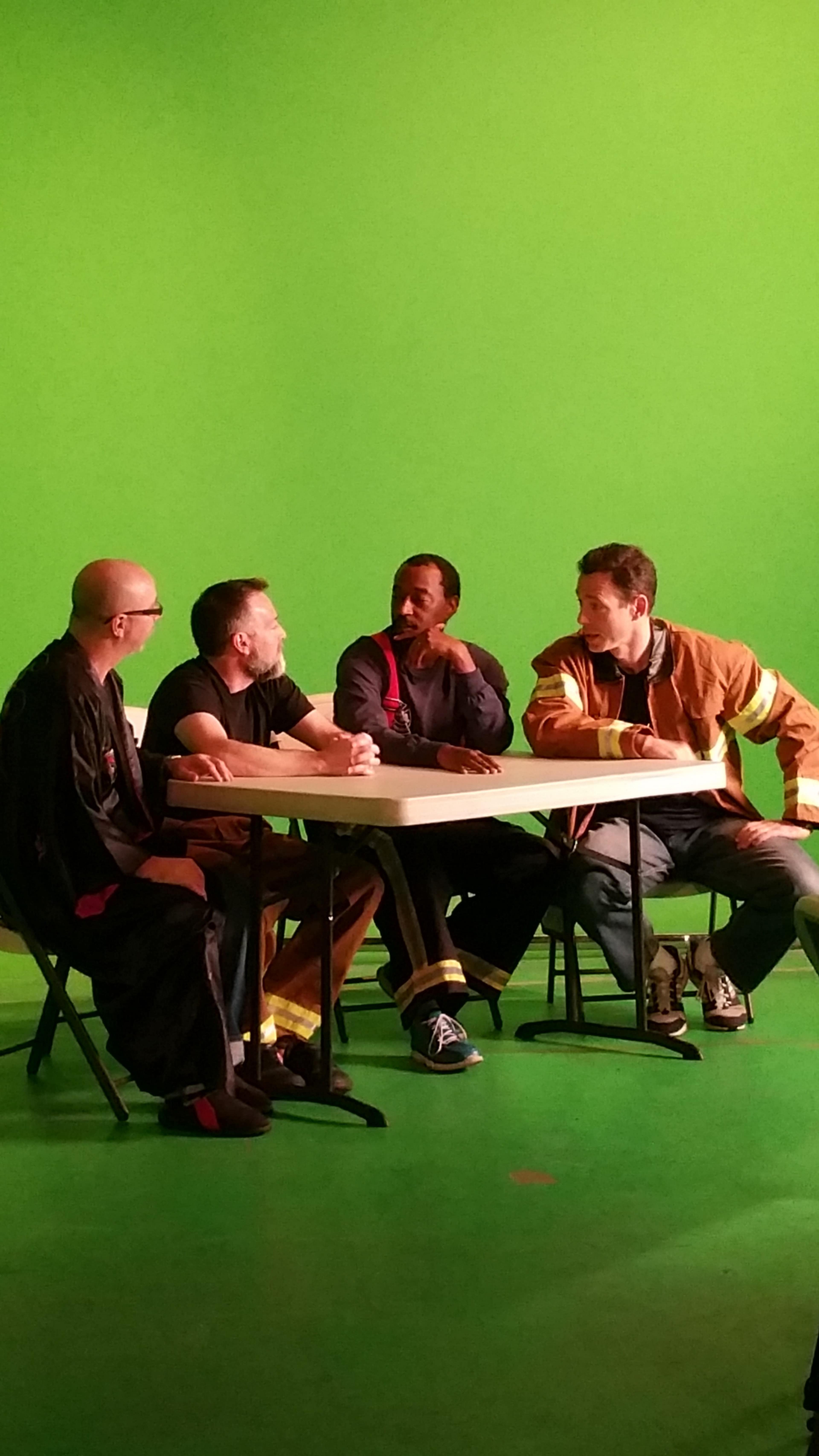 Four men sit around a table in a green screen studio, engaged in conversation.