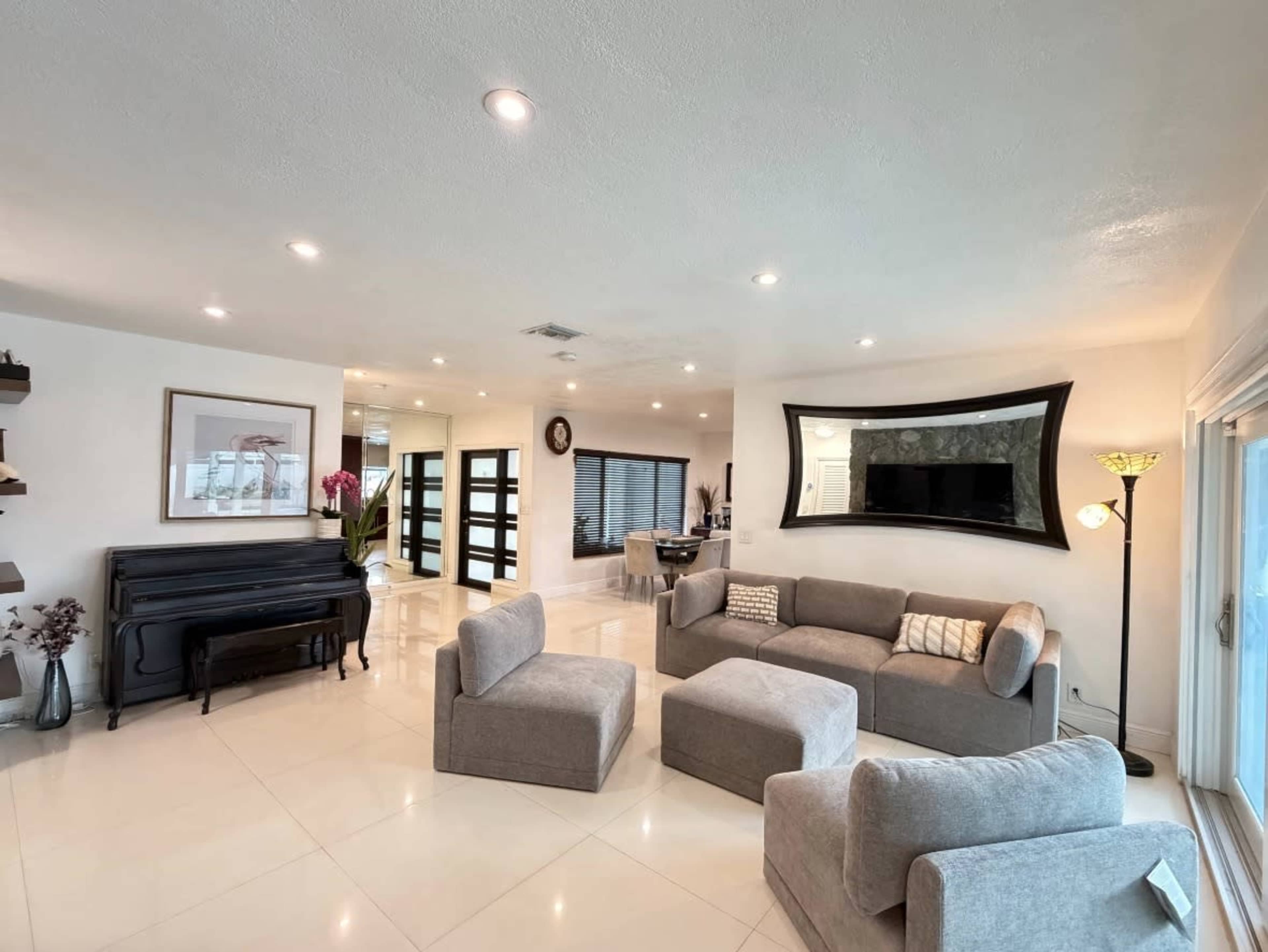 The image shows a modern living room with a piano, various seating arrangements, and large windows allowing natural light to enter.