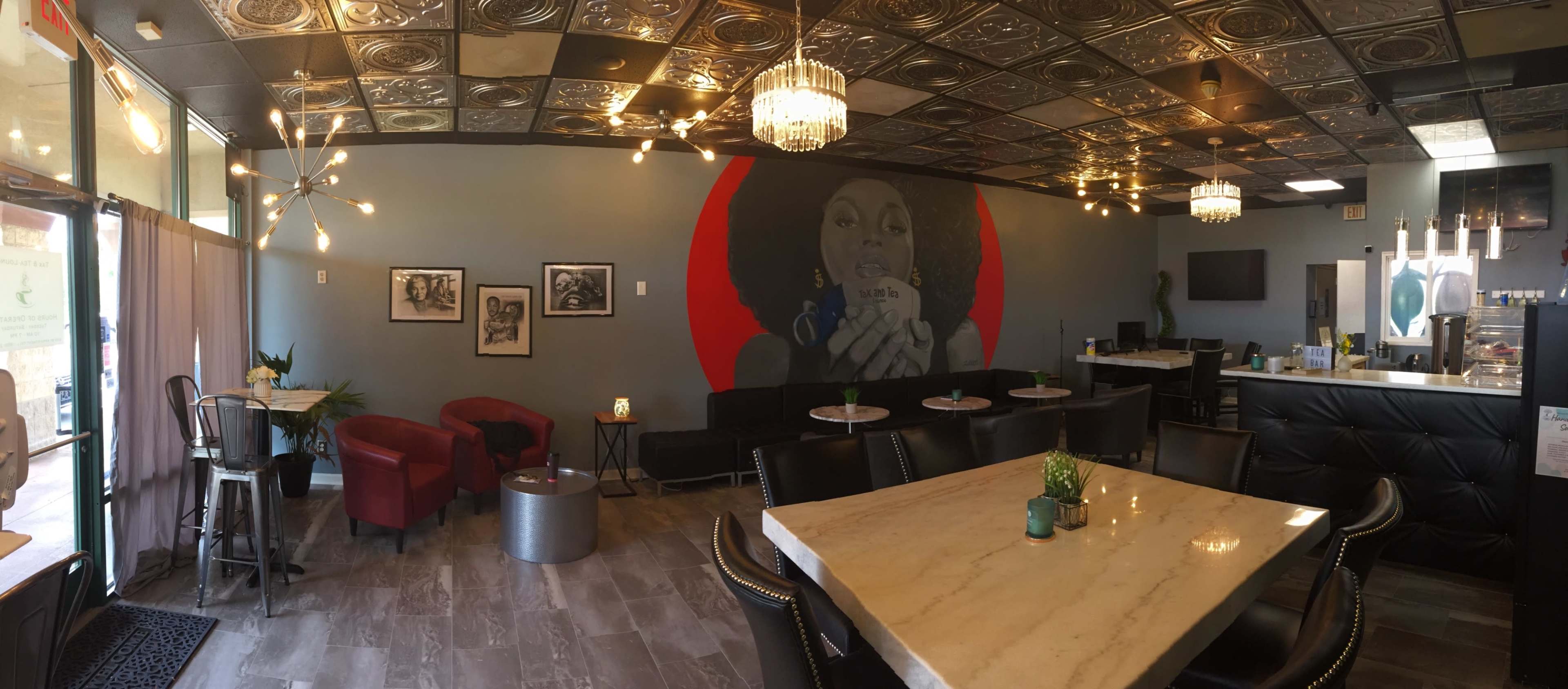A modern café with a large mural on one wall, featuring a woman holding a cup, and a seating area consisting of a marble table and red armchairs.