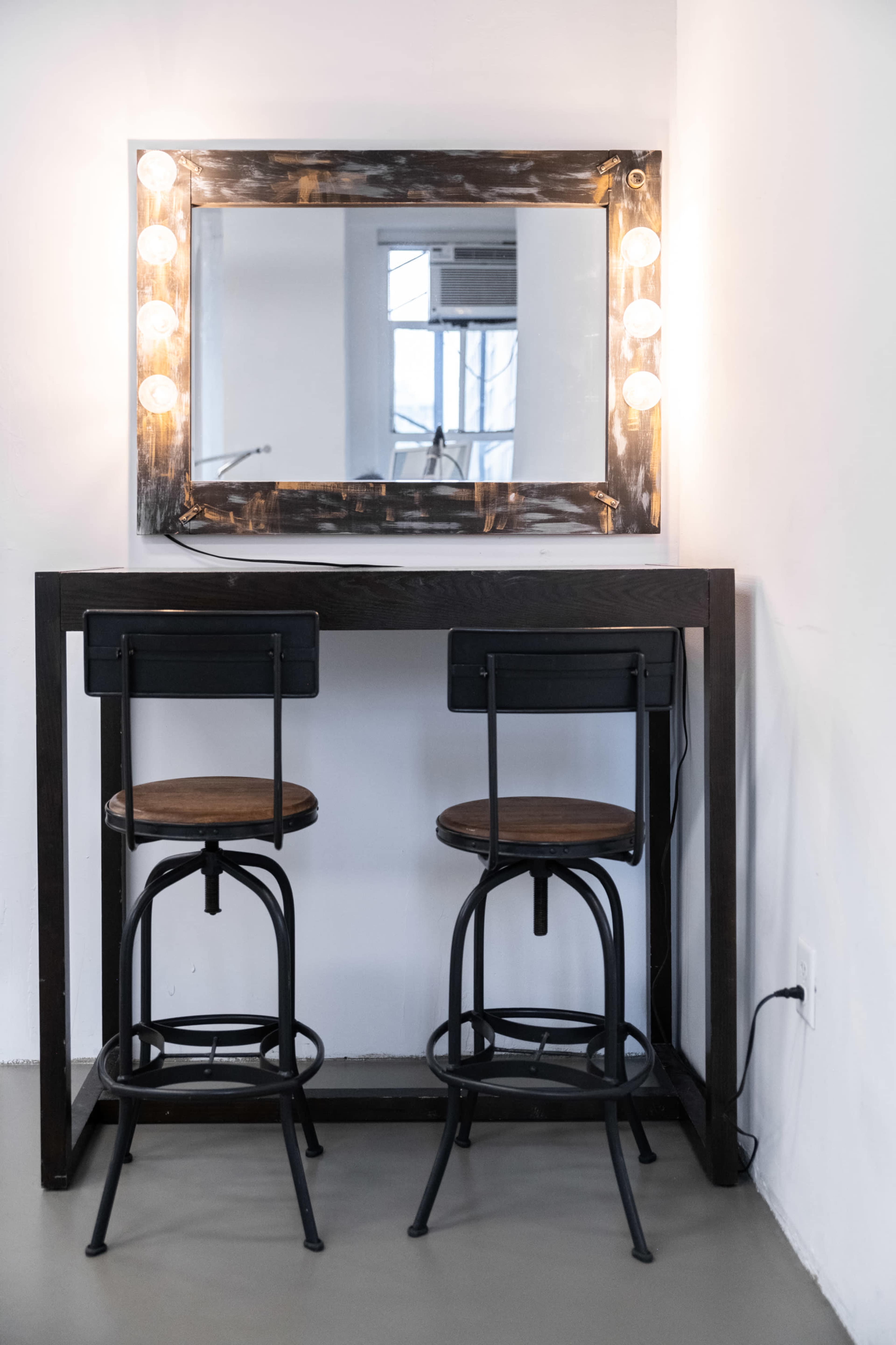 A minimalist vanity setup features a dark wooden table with two metal-framed stools and a large illuminated mirror on the wall.