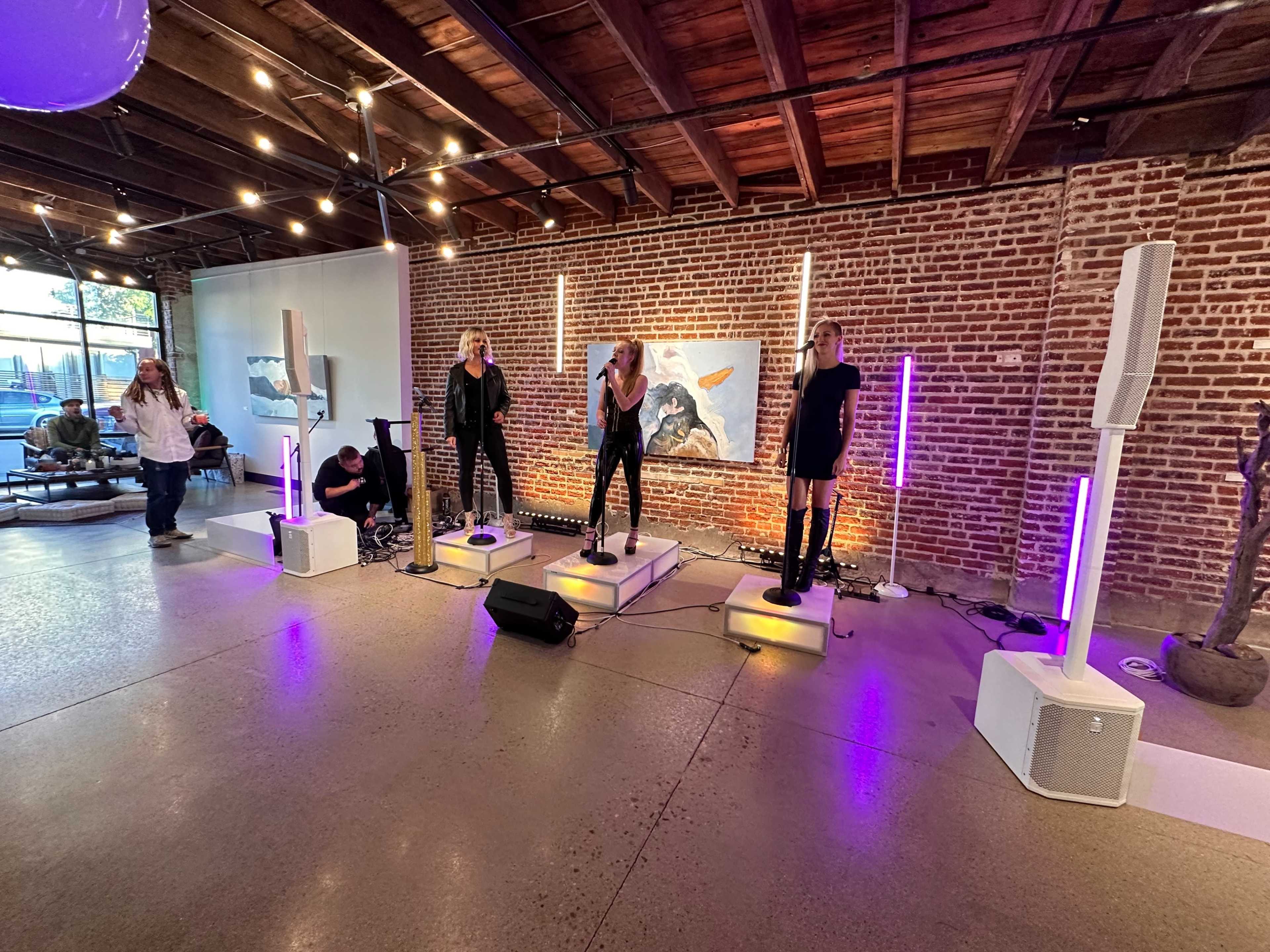Four performers sing on elevated platforms with lights in a loft-style venue featuring exposed brick walls.