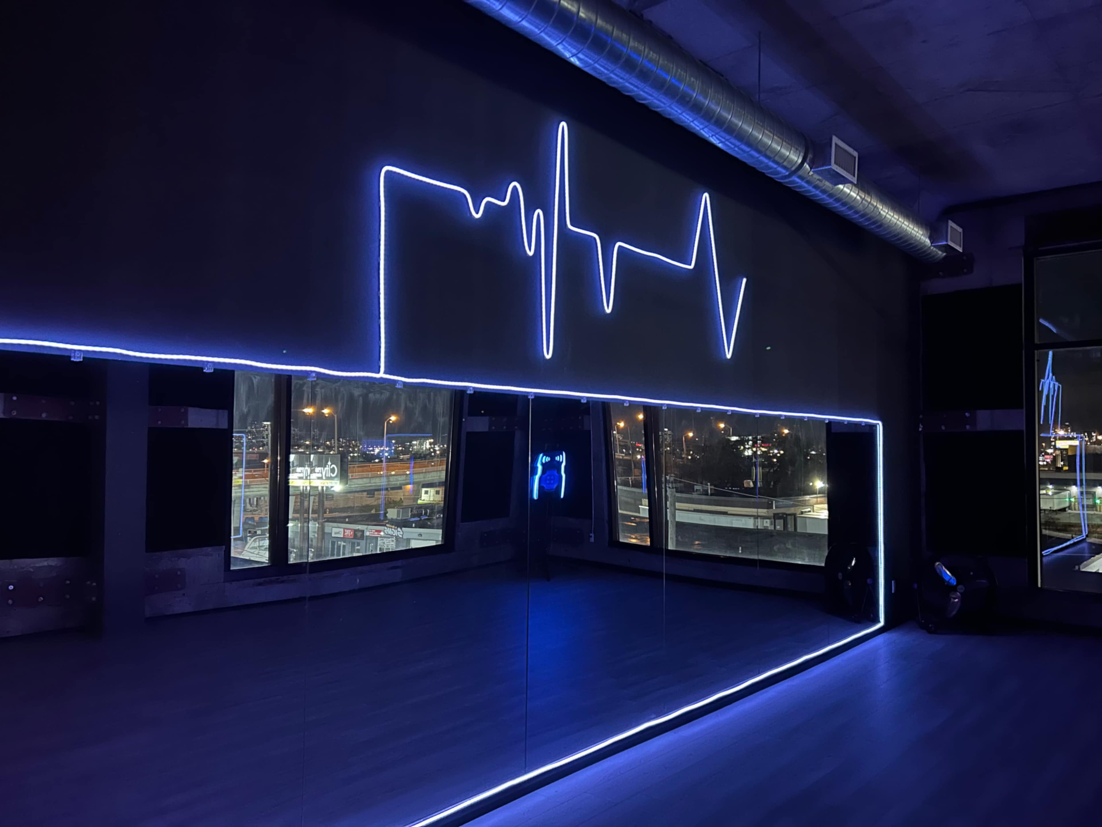 The image shows a modern fitness studio featuring a large mirror and a neon light display designed to resemble a heartbeat line, set against dark walls.