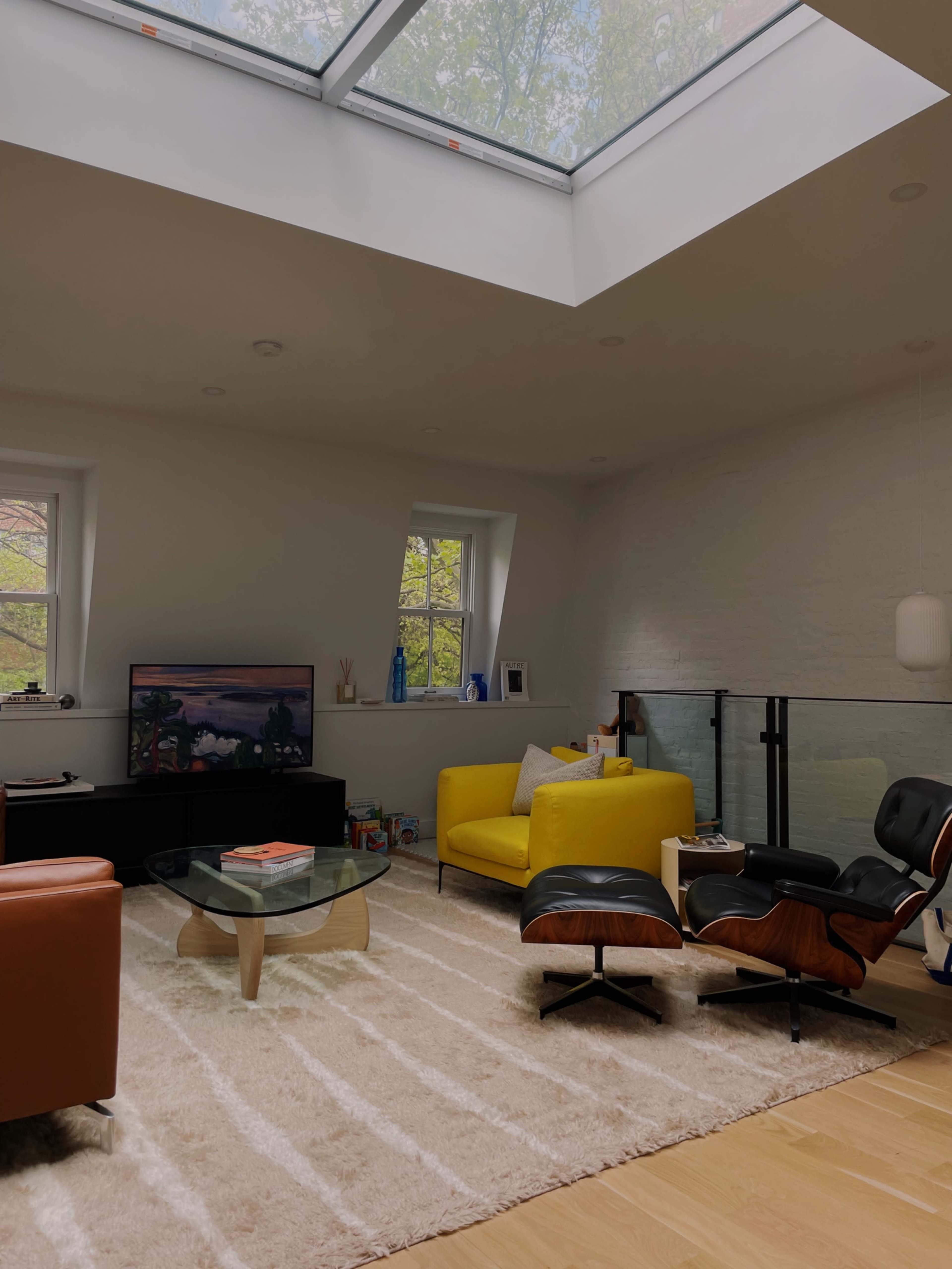 The image shows a modern living room with a yellow couch, an orange armchair, a coffee table, and a television under a skylight.