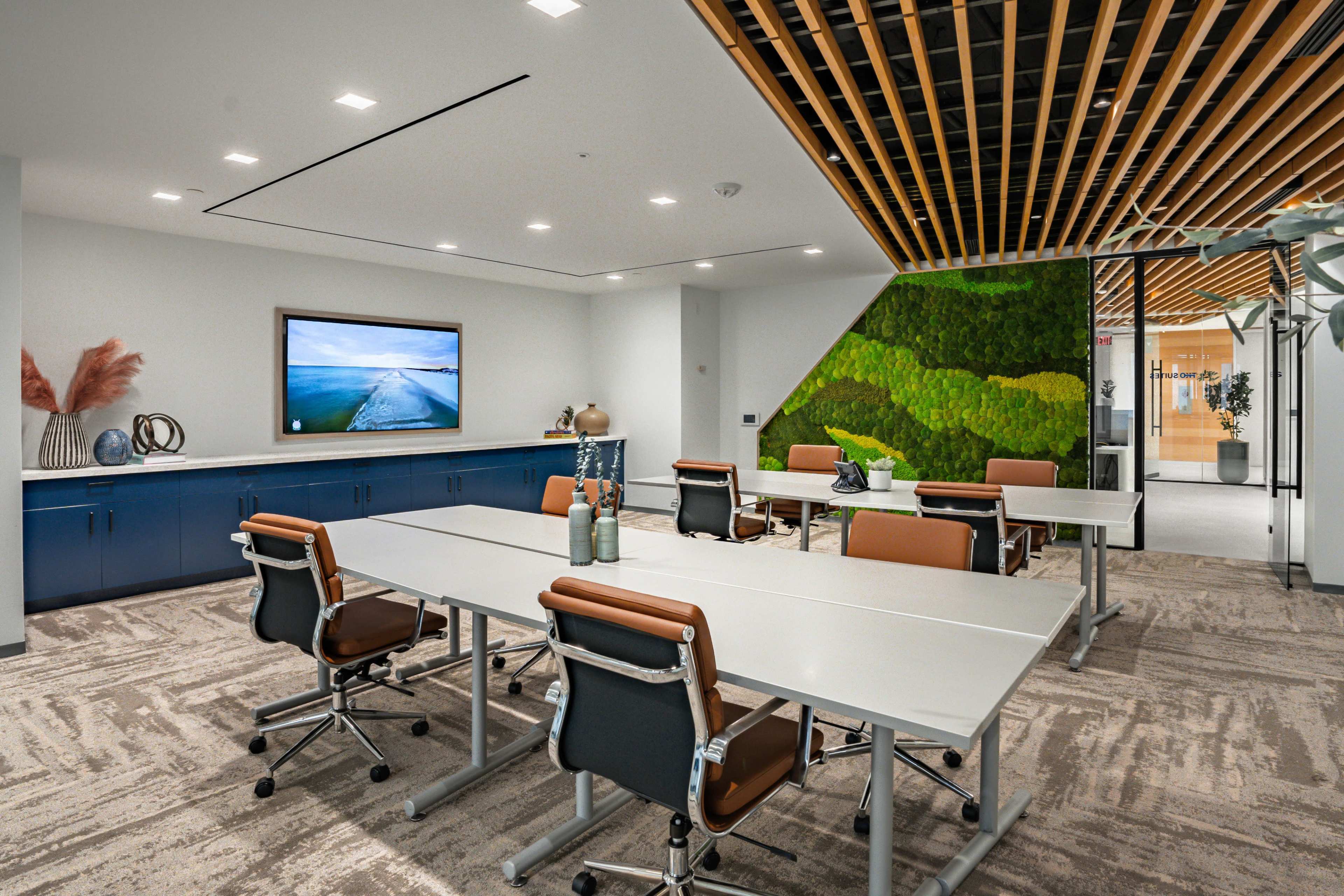 The image shows a modern conference room with several tables and chairs, a large television screen on the wall, and a green wall feature.