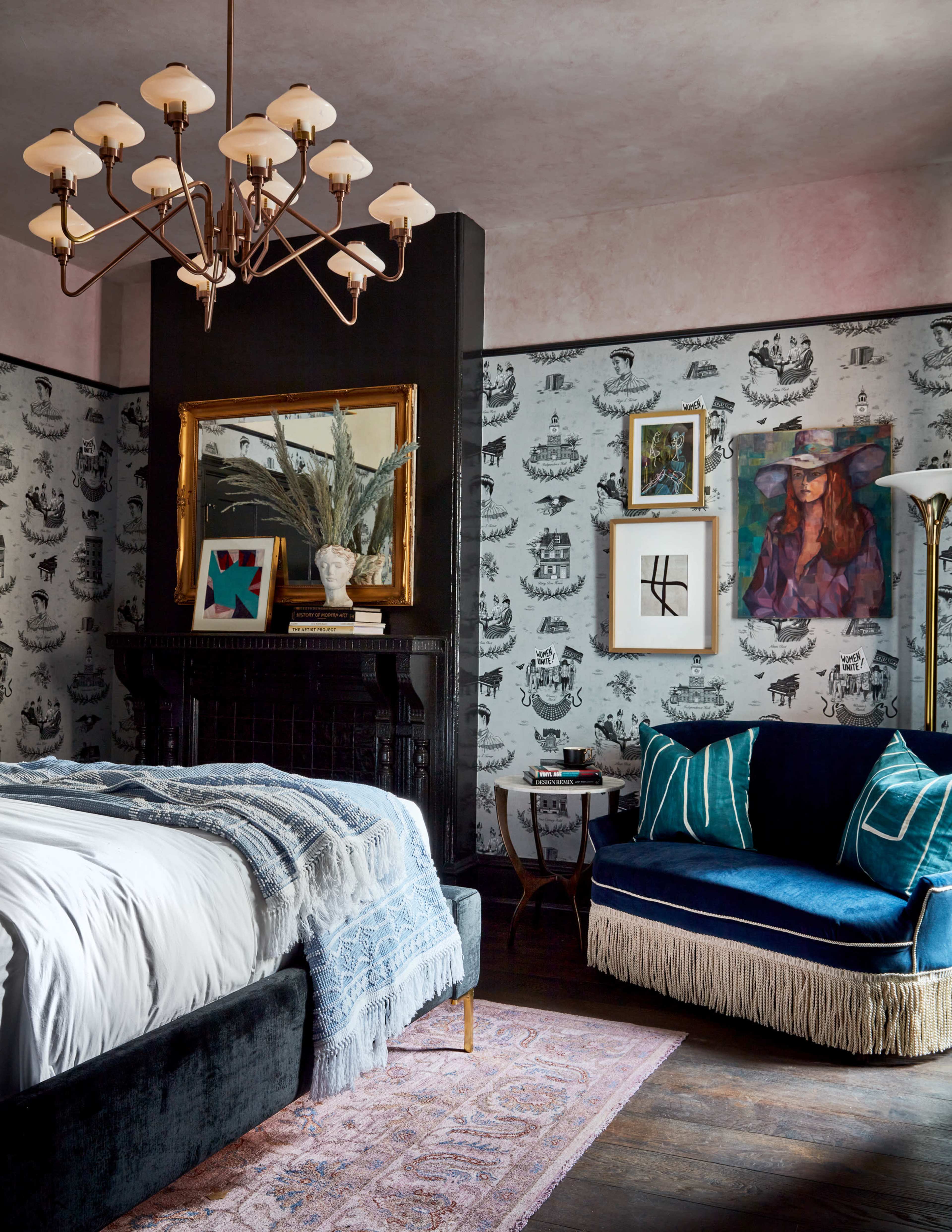 The image depicts a stylish bedroom featuring a black accent wall with decorative wallpaper, a bed with a light-colored blanket, a blue velvet sofa with fringes, and various framed artworks on the walls.