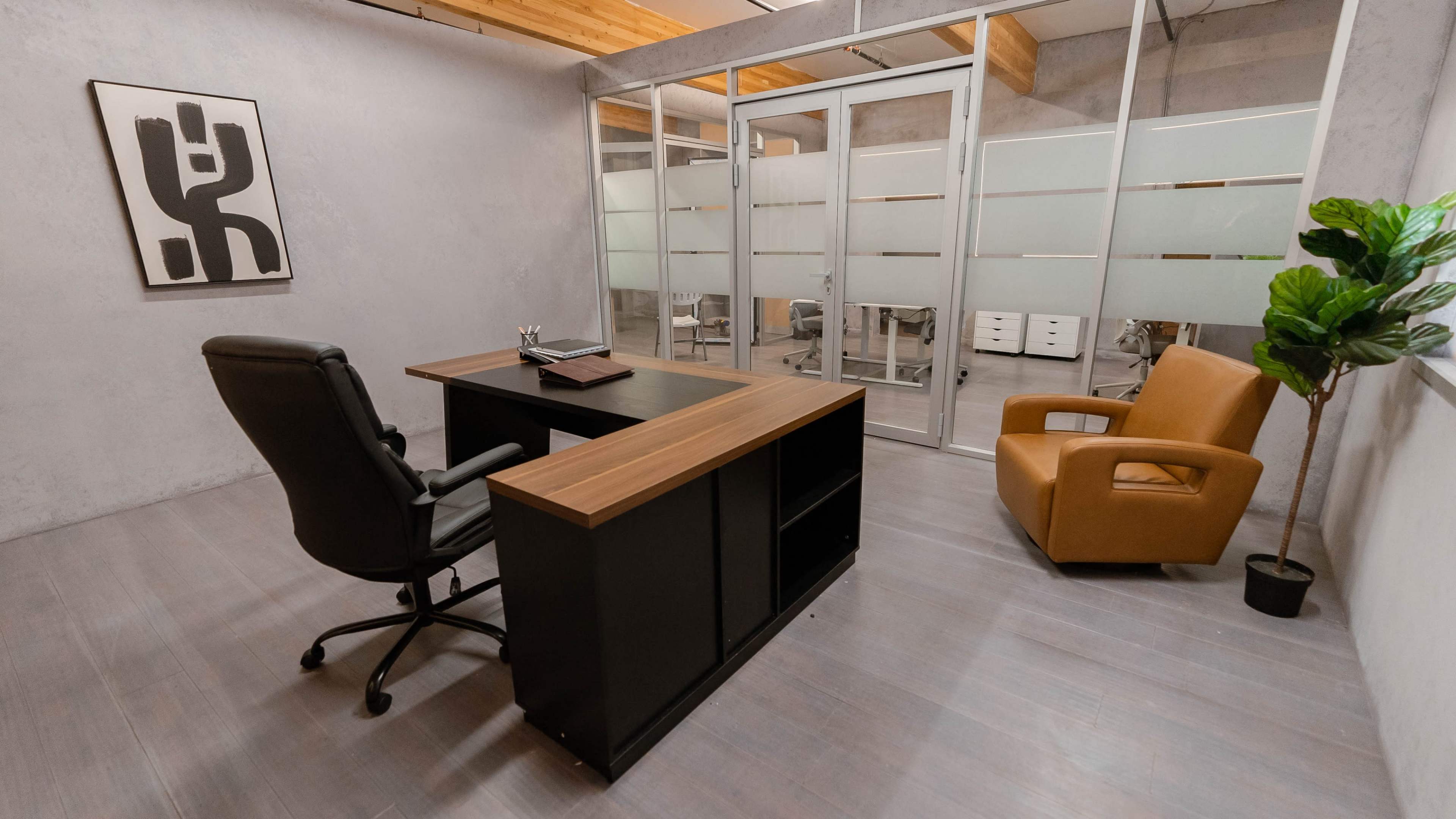 The image shows a modern office space featuring a black desk with a wooden top, a swivel chair, an orange armchair, and a plant in the corner.