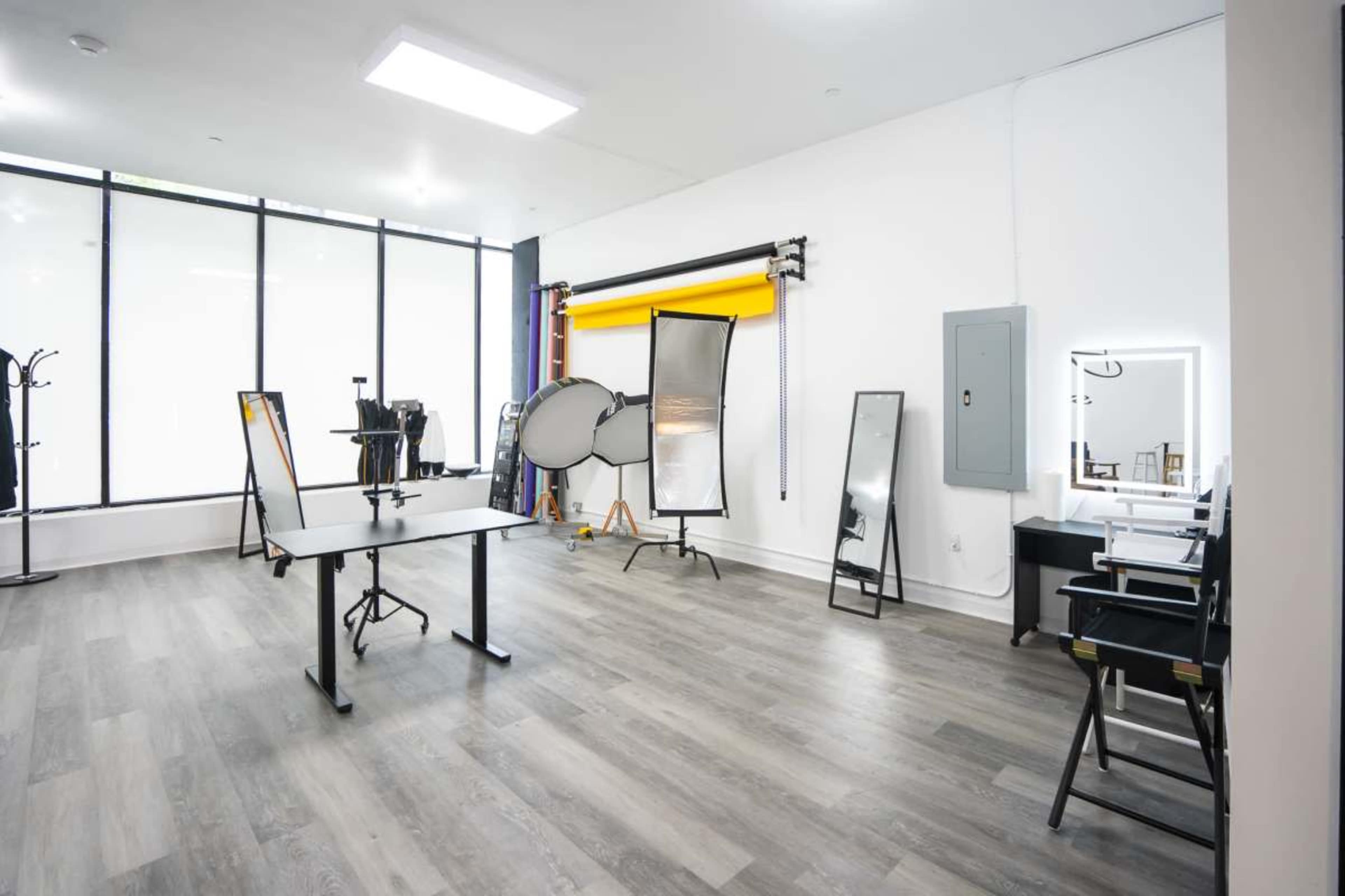 The image shows a bright, spacious photography studio featuring large windows, a central table, multiple mirrors, and backdrop equipment.