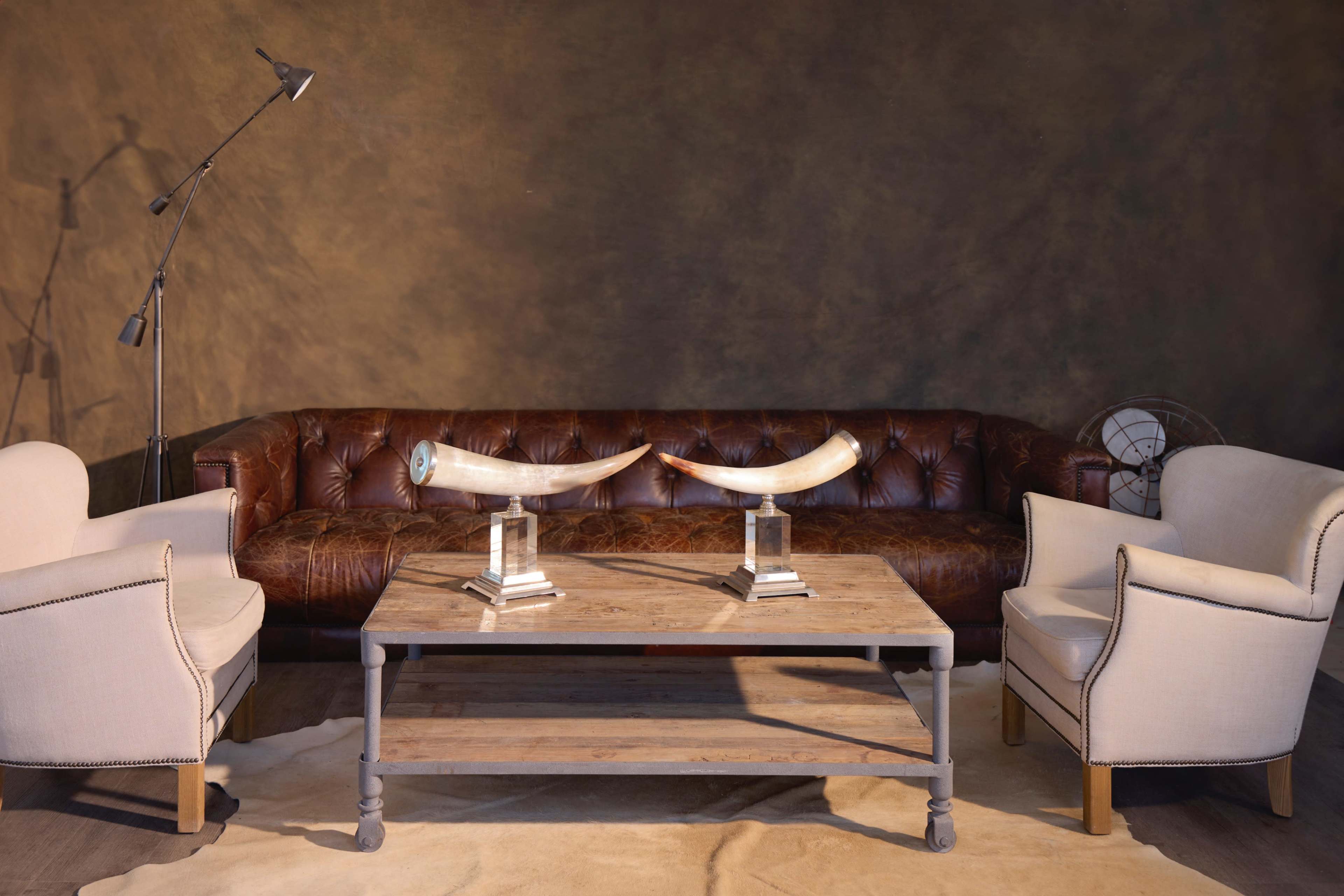 A brown leather chesterfield sofa paired with two light-colored armchairs, centered around a wooden coffee table with decorative tusks displayed on glass stands, set against a textured brown wall.