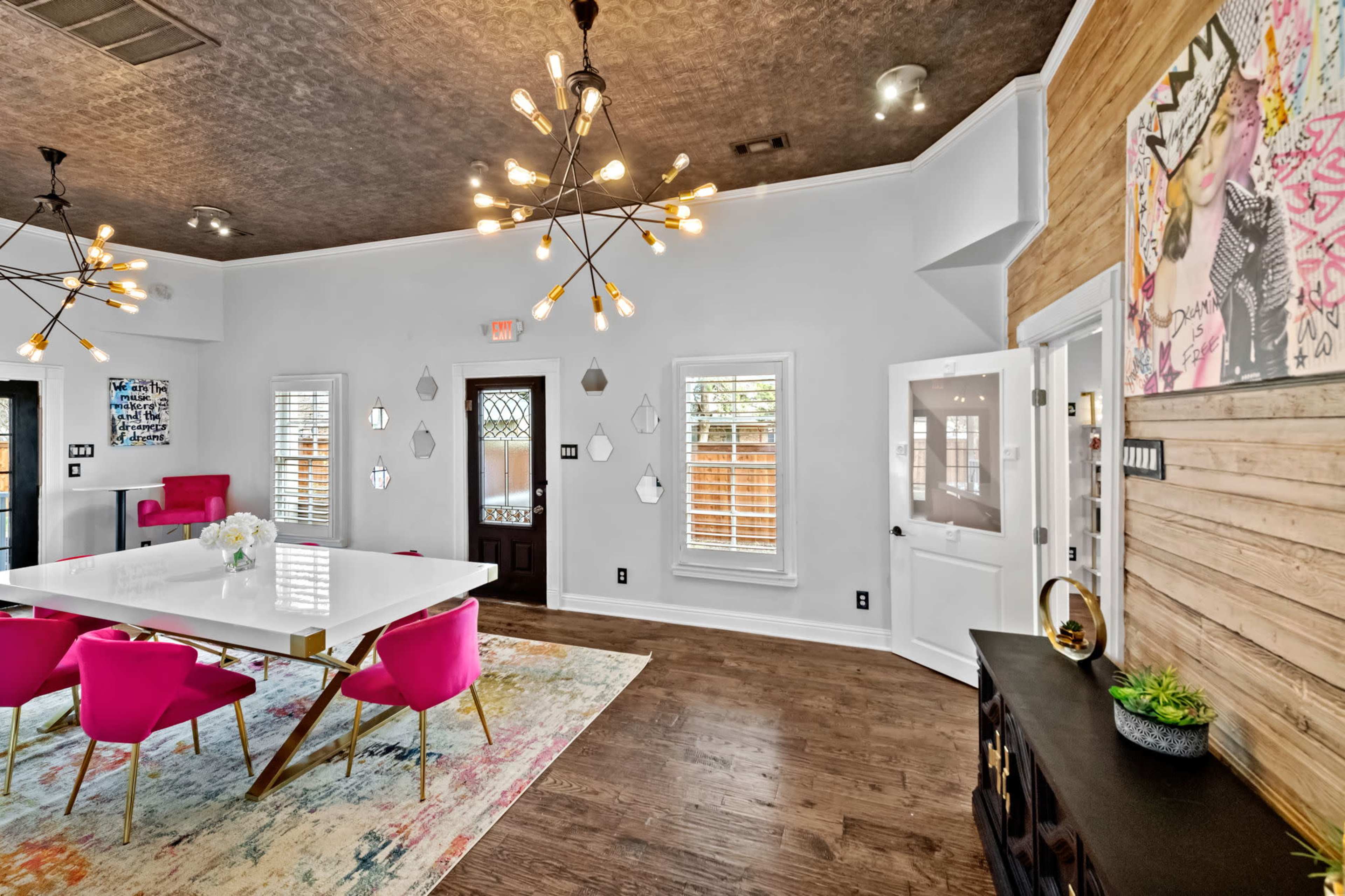 The interior features a dining area with a large white table surrounded by pink chairs, illuminated by modern light fixtures, against a backdrop of wood and decorative wall art.