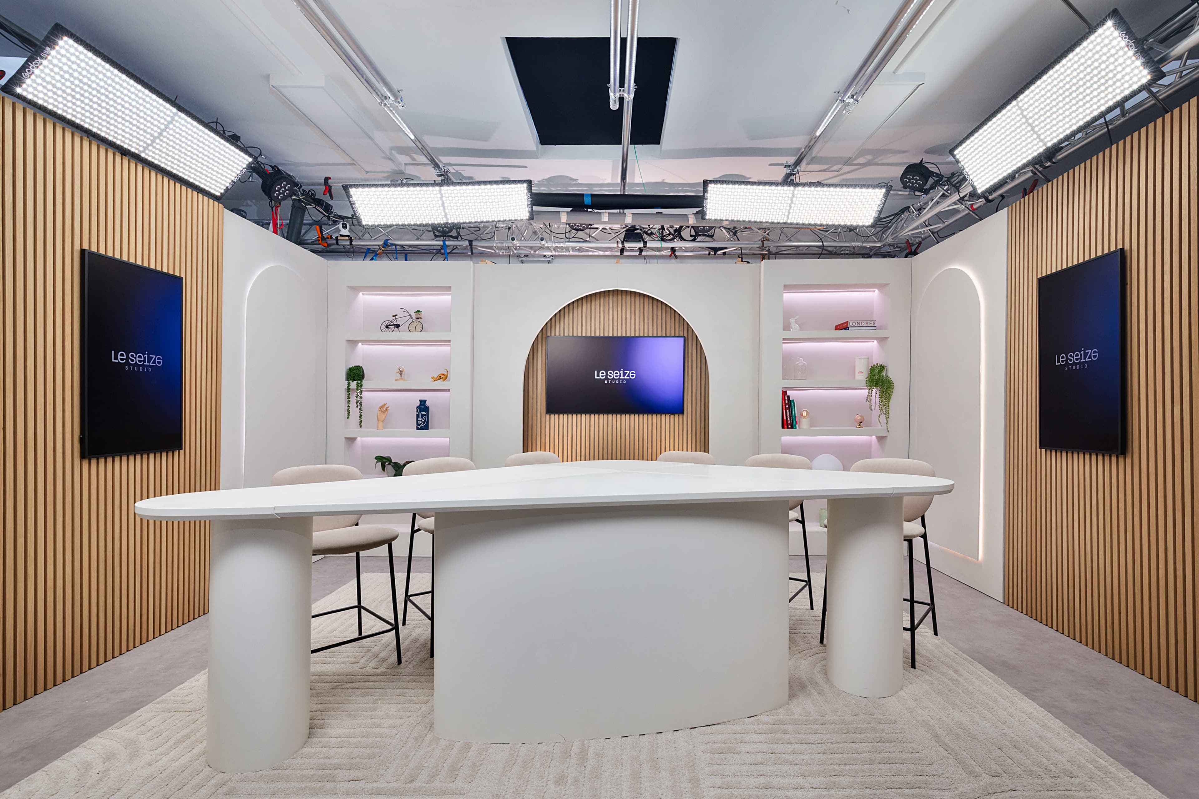 The image shows a modern studio setup with a curved white table, bar stools, and two large screens on the walls, surrounded by wooden paneling and shelves.