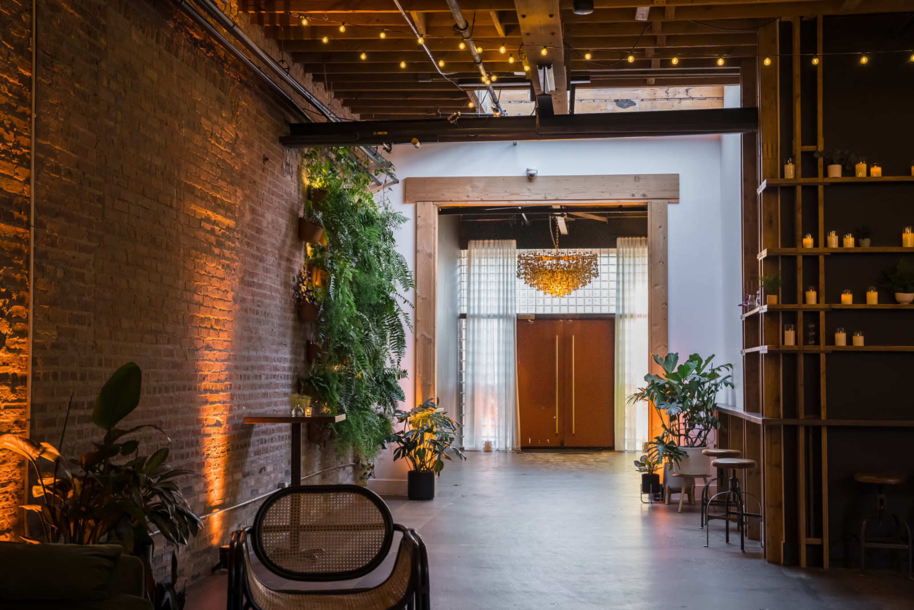 The image shows a spacious indoor area with brick walls, wooden beams, plants, and soft lighting, leading to a pair of double doors at the far end.