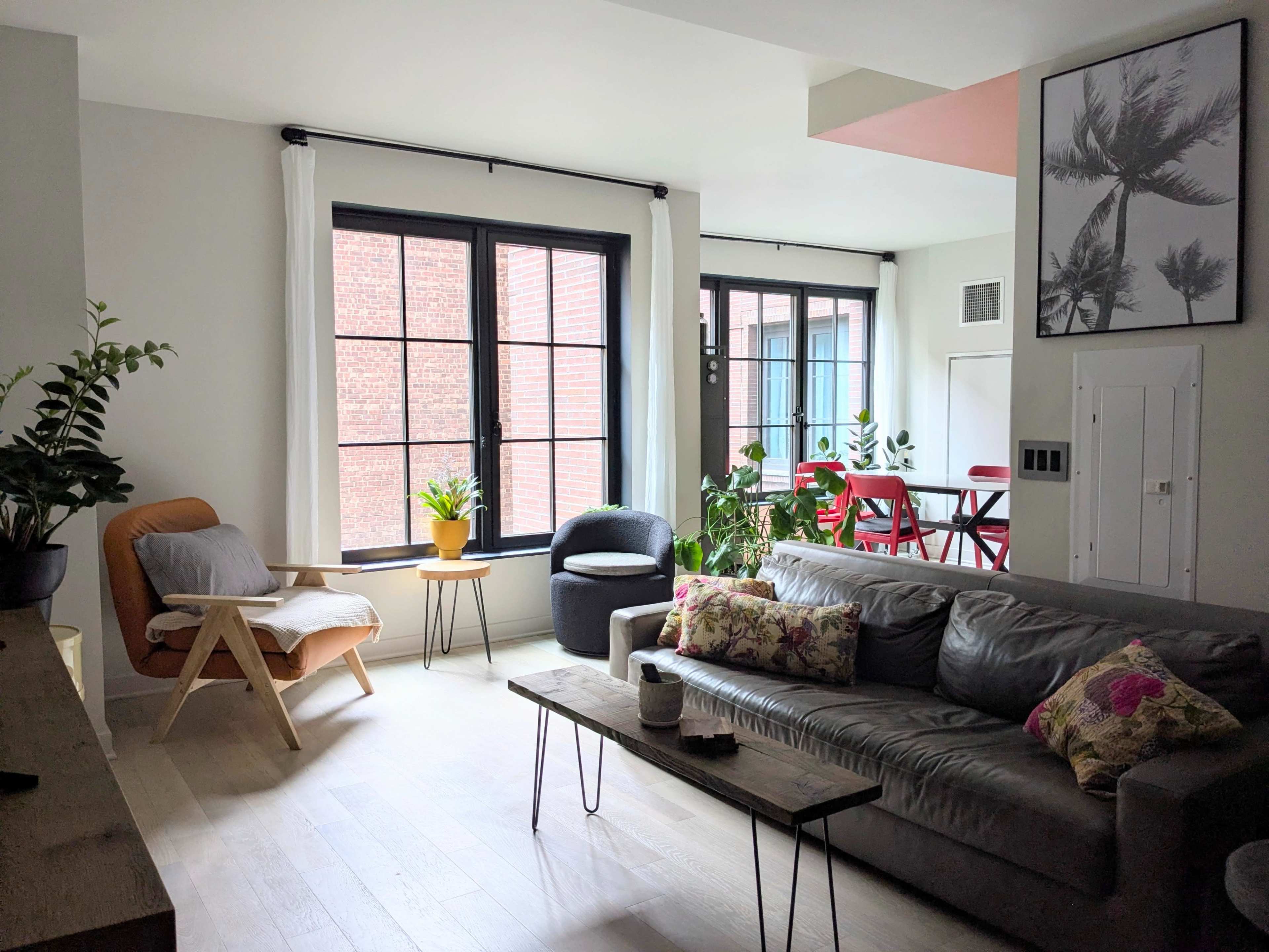 The image shows a modern living room with a gray sofa, a wooden coffee table, large windows letting in natural light, and plants decorating the space.