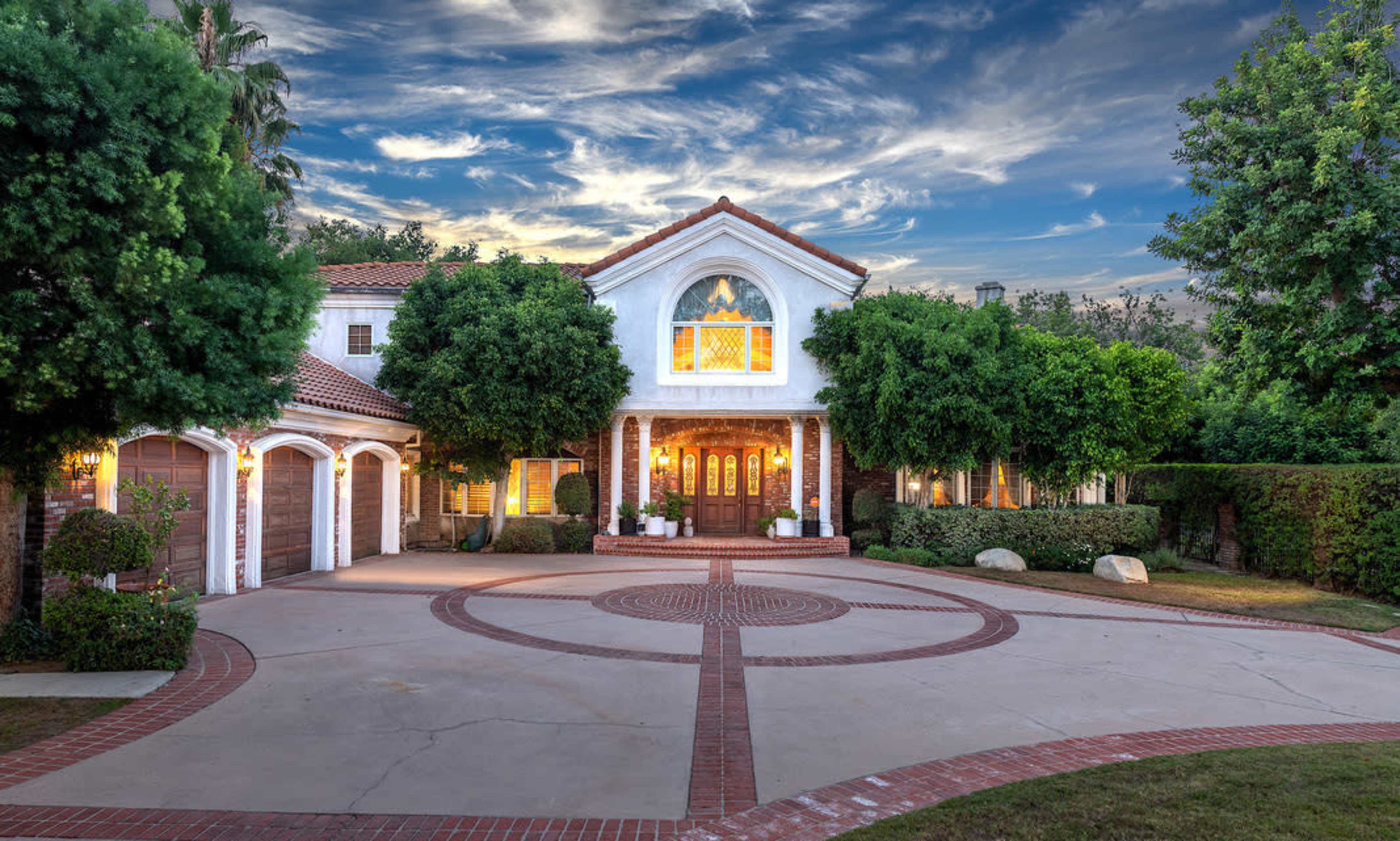 The image shows a large, upscale home with a circular driveway, landscaped greenery, and a blue sky with clouds in the background.