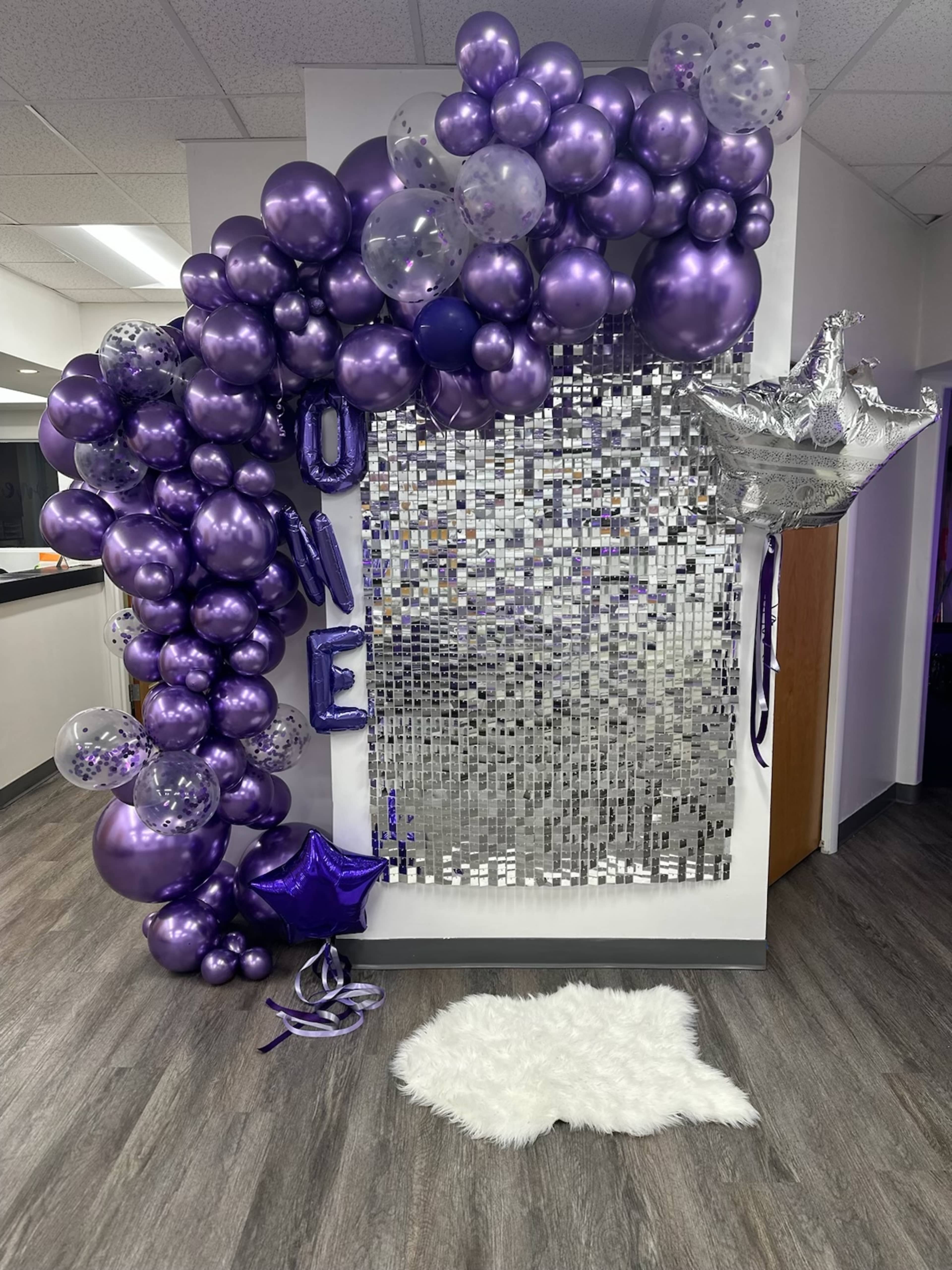 A decorative display featuring a cluster of purple balloons and a silver backdrop, along with a star-shaped balloon and a fluffy white rug on the floor.