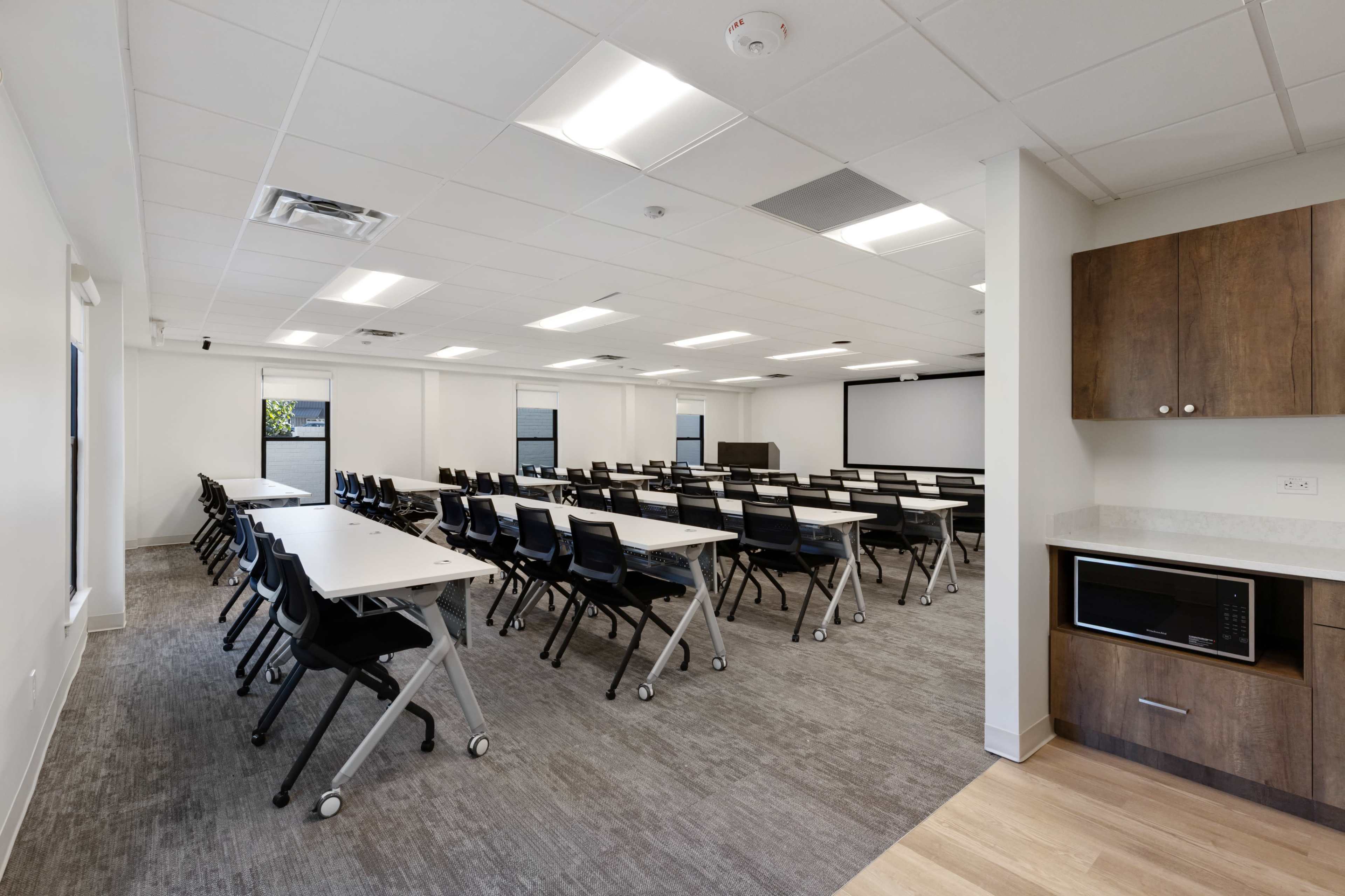 The image shows a modern conference room with rows of tables and chairs arranged facing a projection screen, featuring a kitchenette area in the corner.