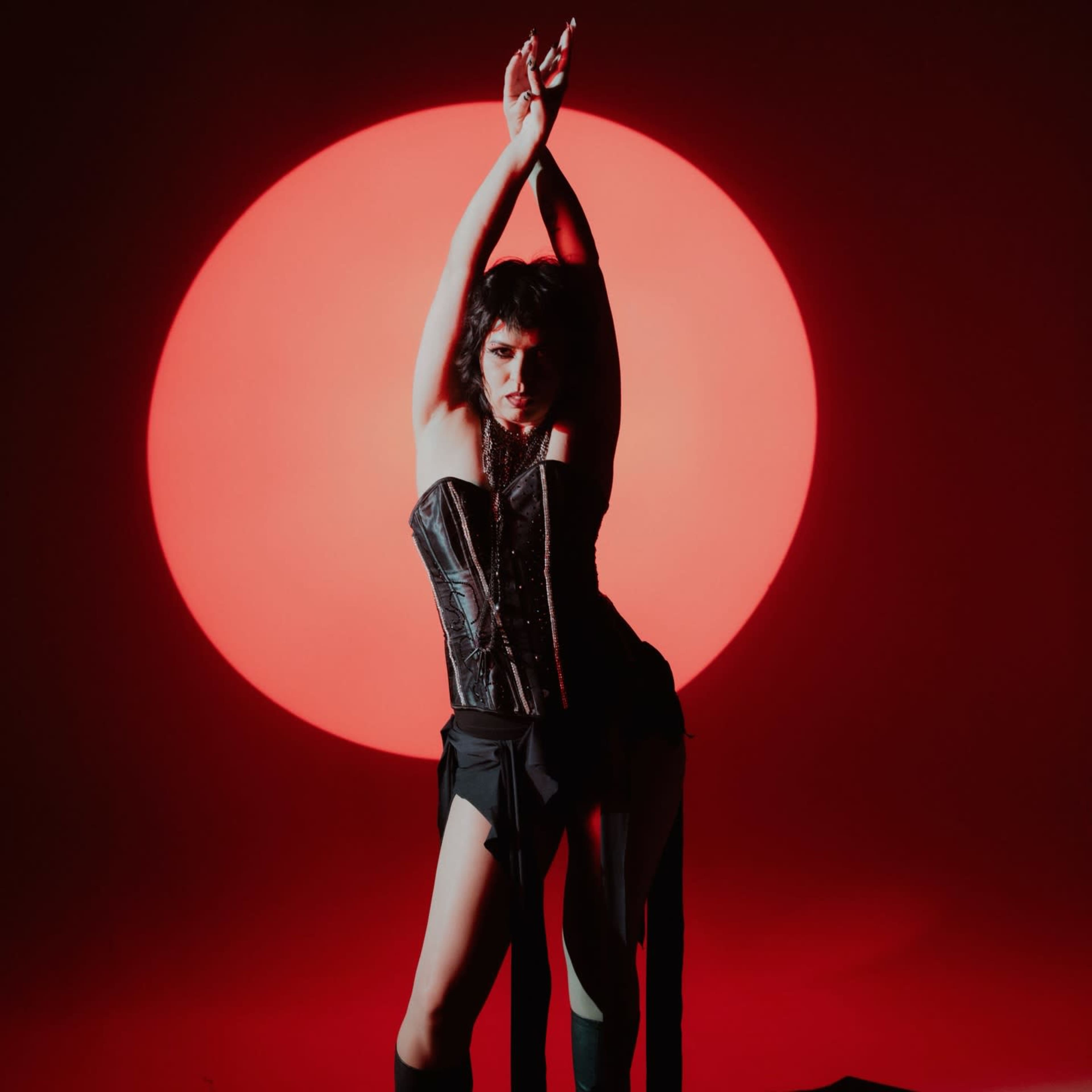 A person poses dramatically in front of a large red circular backdrop, wearing a black corset and black attire.