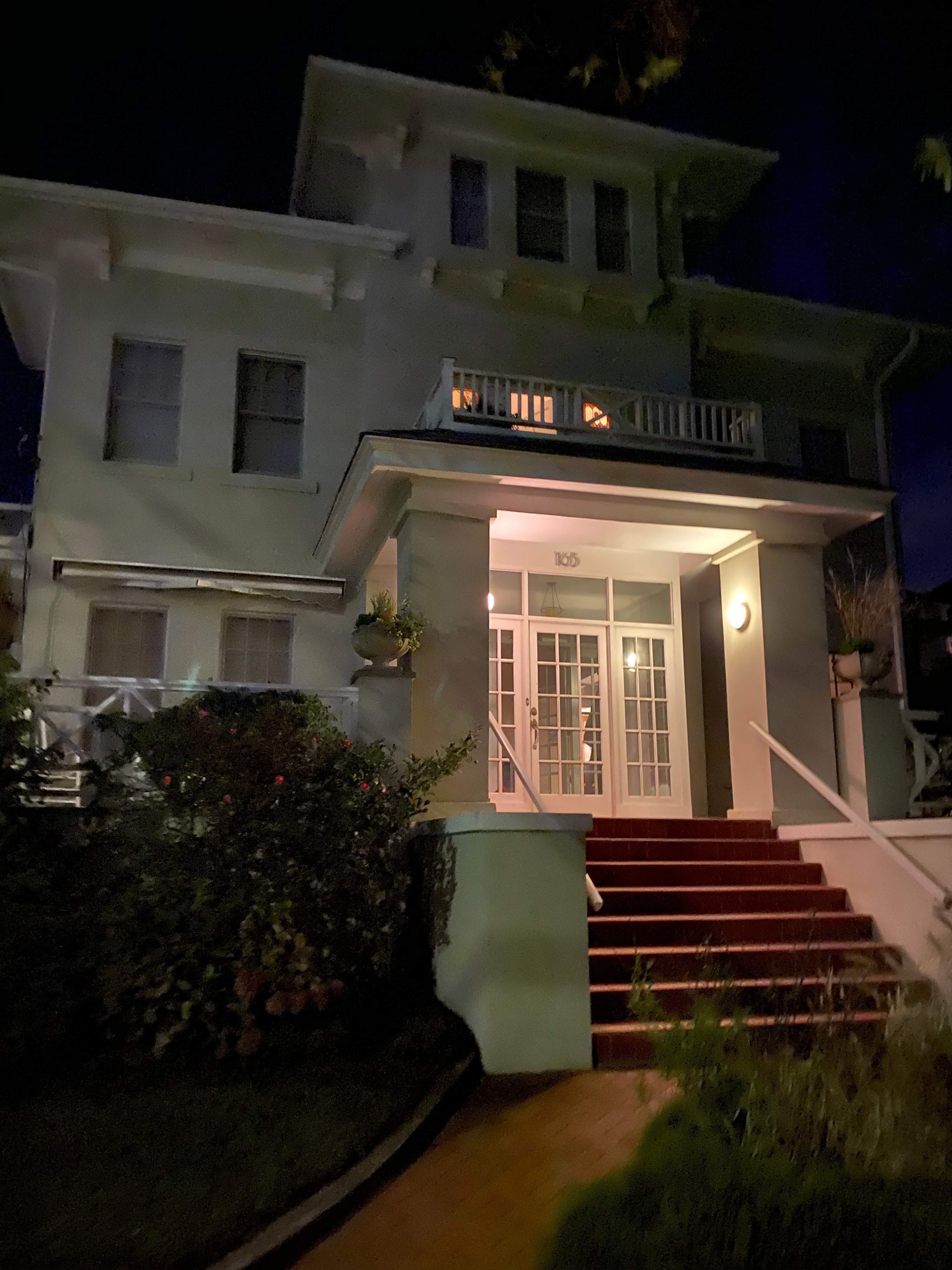 A three-story, white-painted house is illuminated from within, showcasing multiple windows and a red carpet leading up to the entryway at night.
