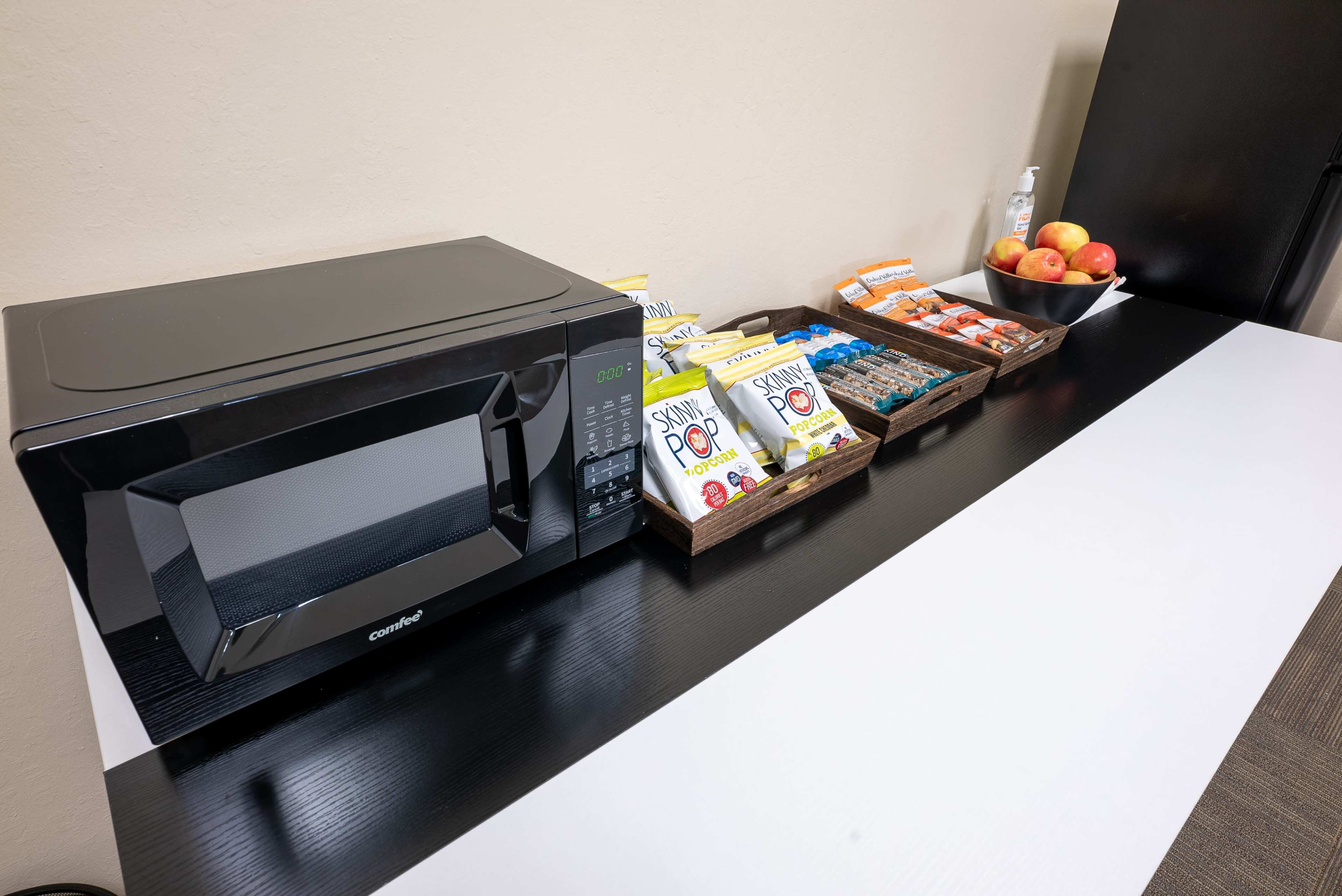 A black microwave is placed on a countertop next to various snacks and apples arranged in baskets.