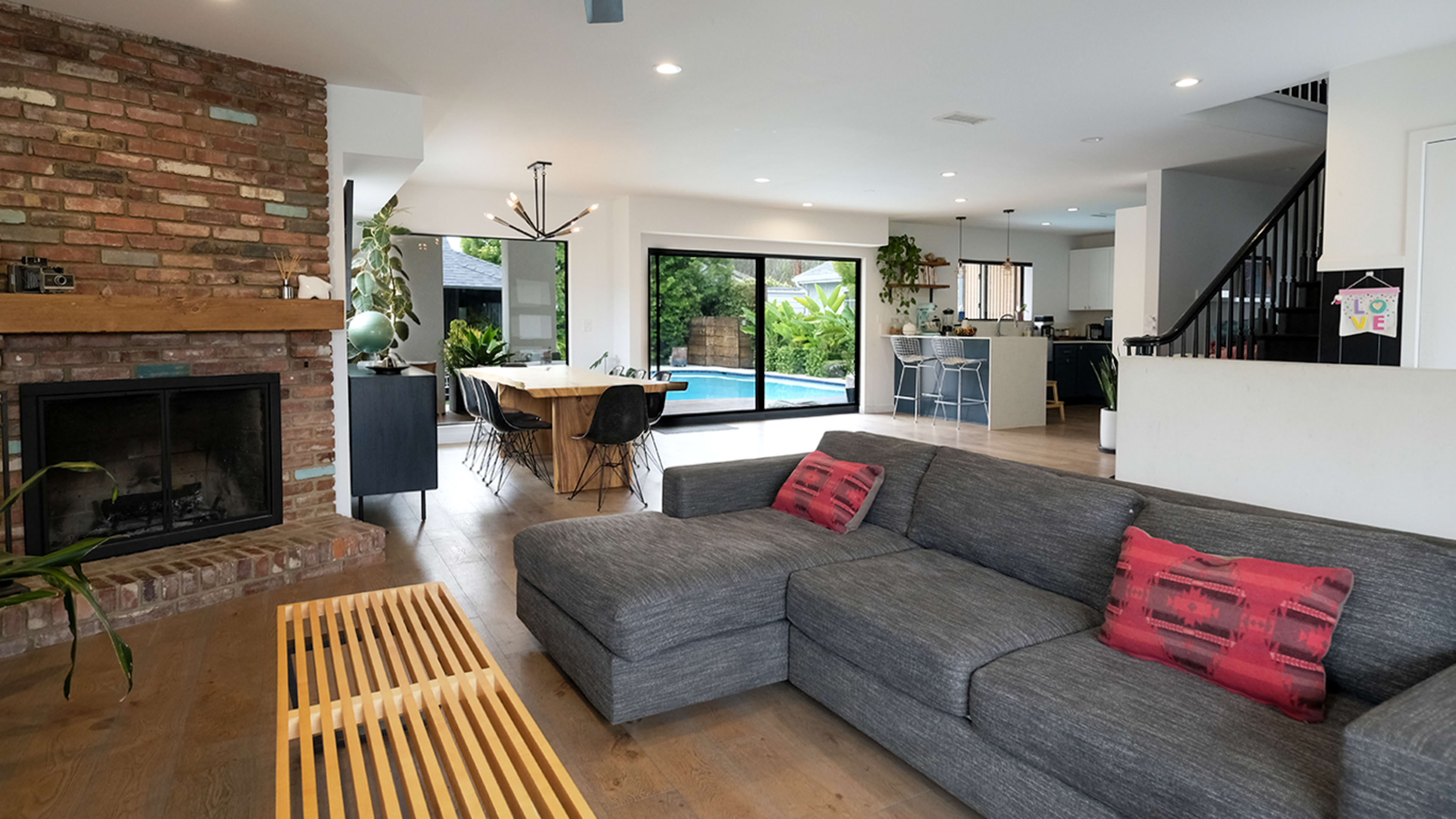 The living area features a gray sectional sofa, a wooden coffee table, and a brick fireplace, with large glass doors leading to a poolside view.