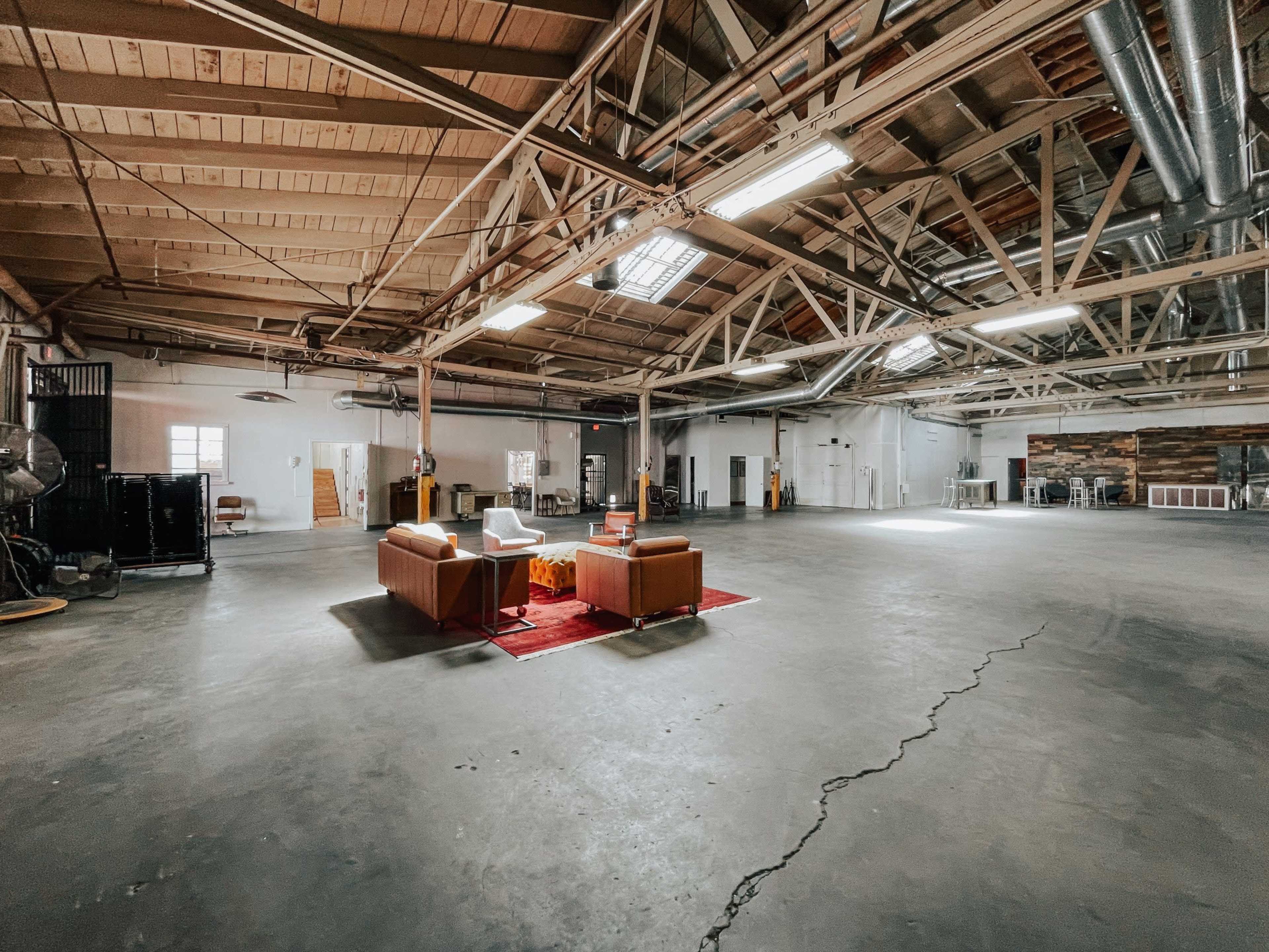The image shows a spacious industrial-style interior with high wooden beams and exposed metal, featuring a minimal seating arrangement on a carpet in the center.