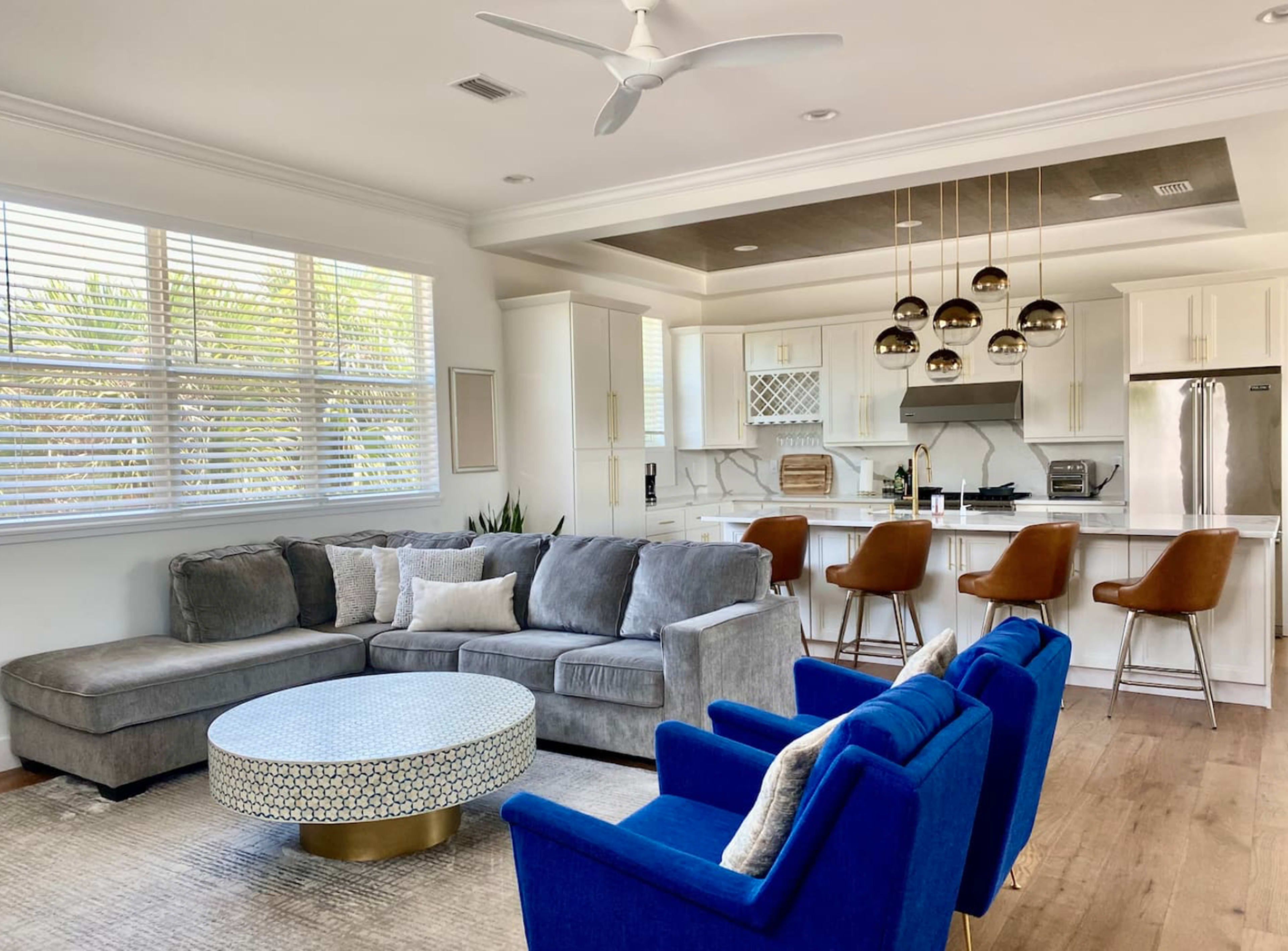A modern living room featuring a gray sectional sofa, a round coffee table, and blue accent chairs, with a kitchen area in the background.