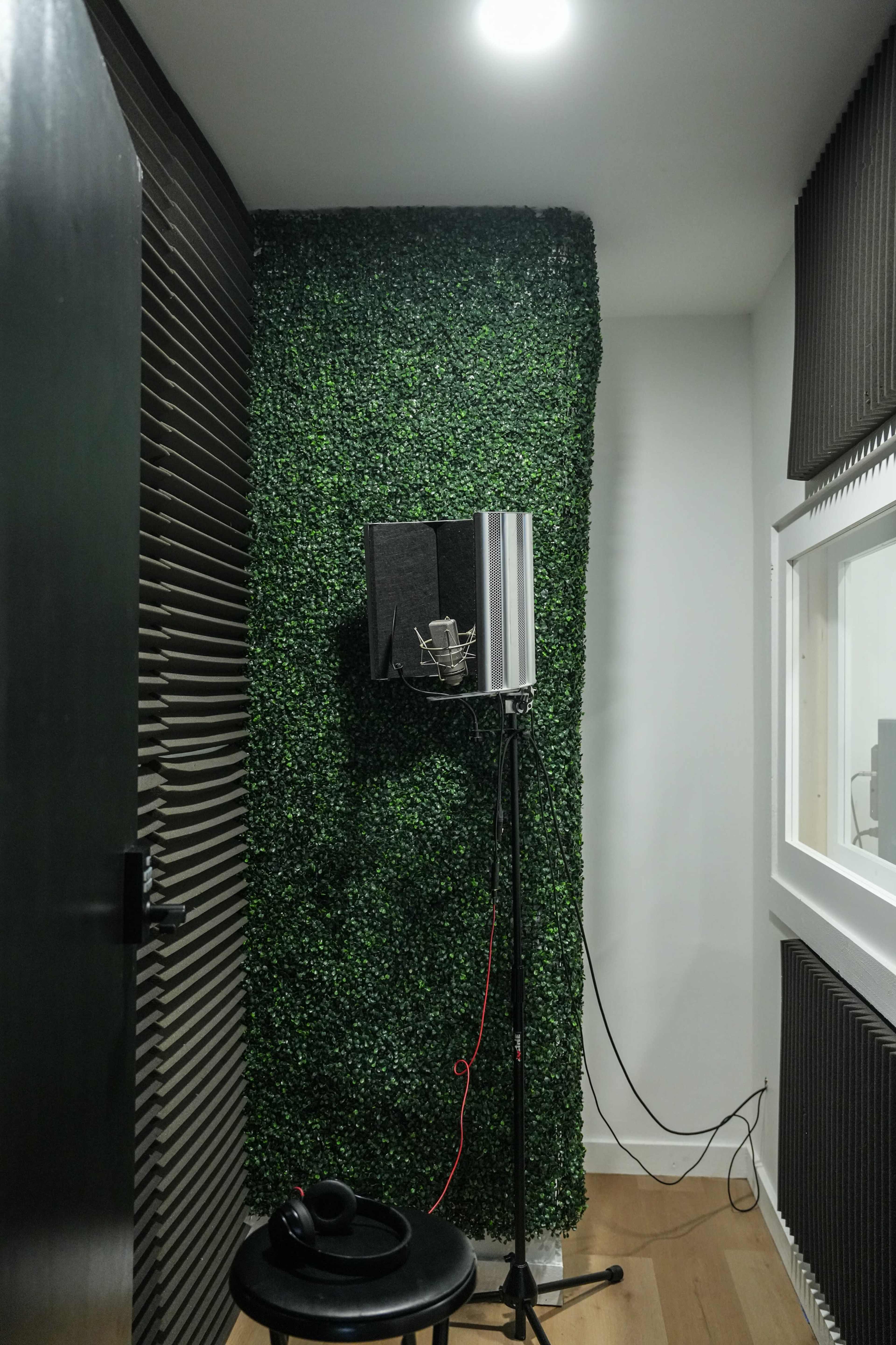 The image shows a small recording studio with a green wall covering, a microphone on a stand, and a stool nearby.