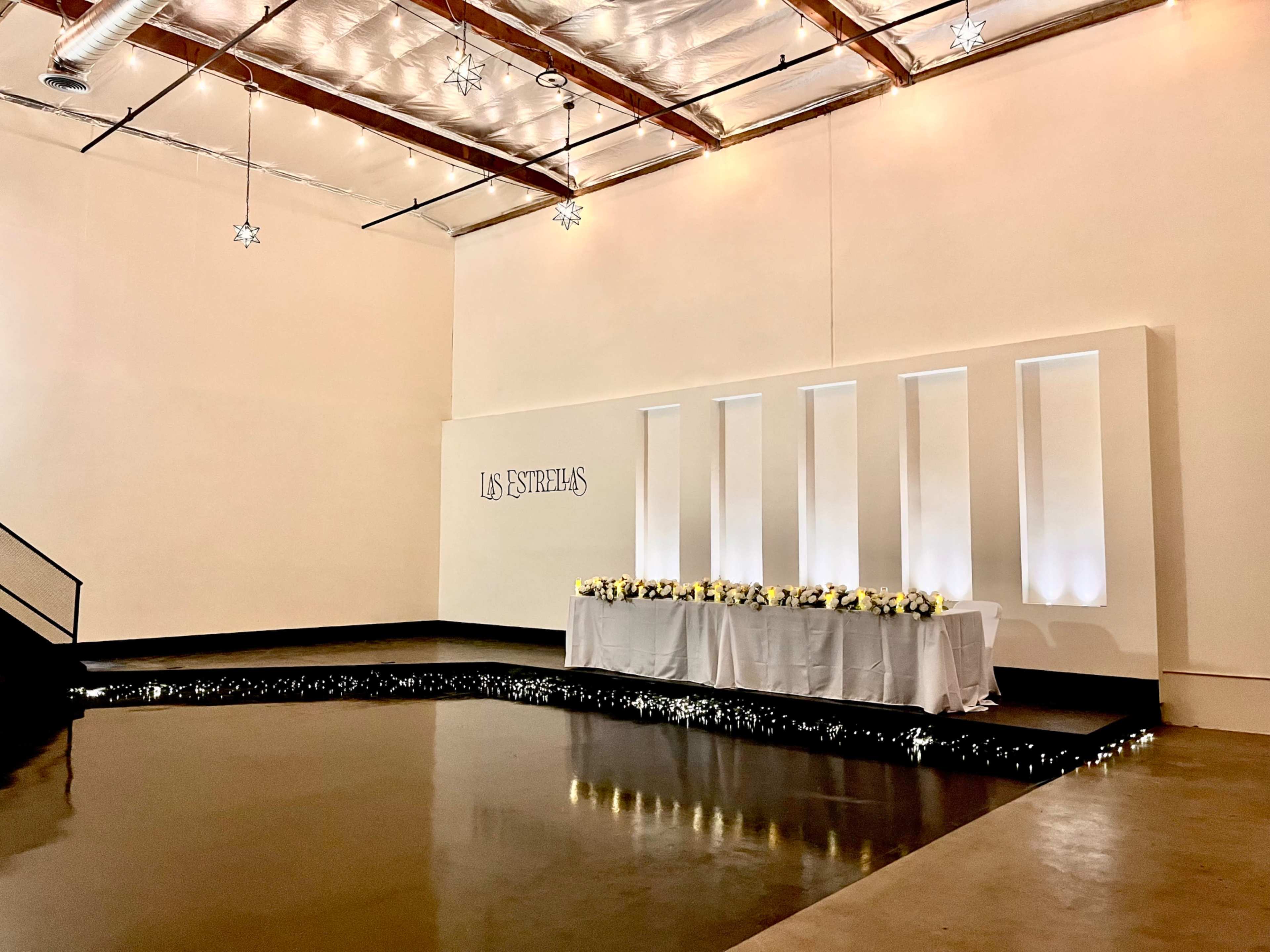 The image shows a spacious, minimalist event venue with a long table adorned with flowers, set against a backdrop of illuminated wall panels and a reflective floor.