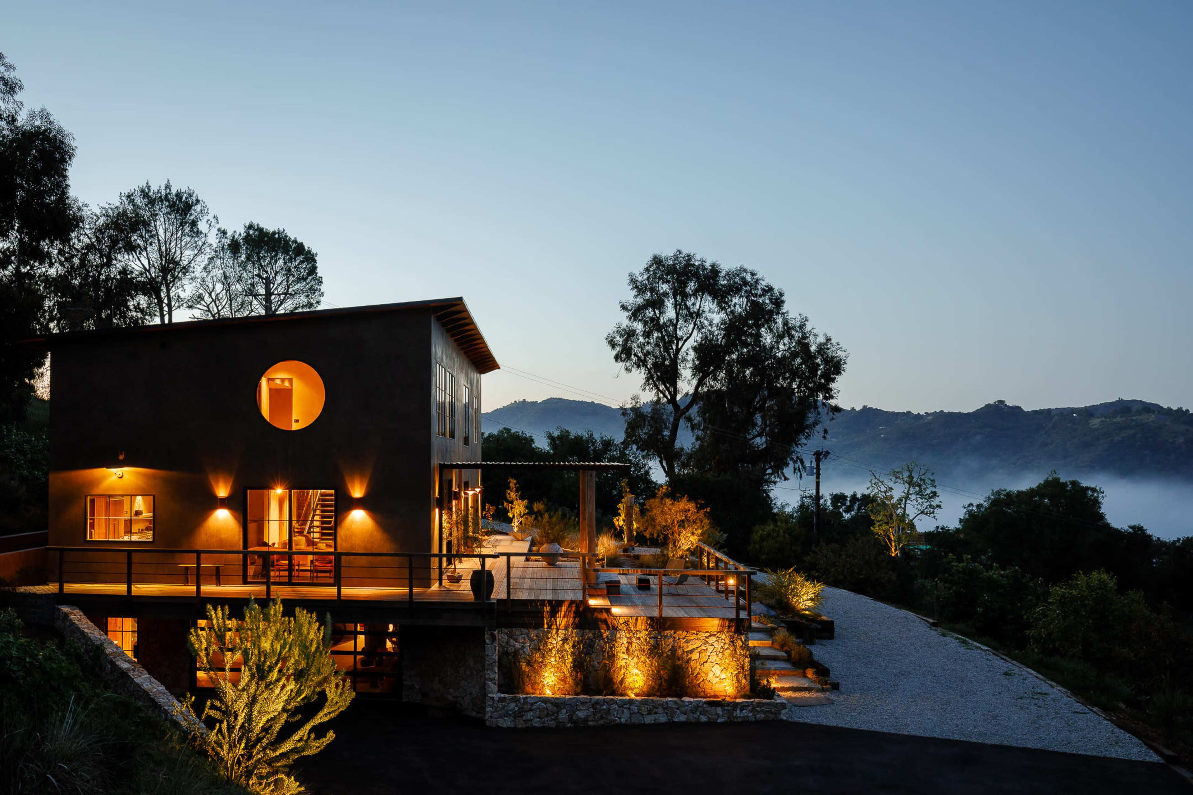 A modern two-story house with exterior lighting sits on a hillside, surrounded by trees and misty mountains at dusk.