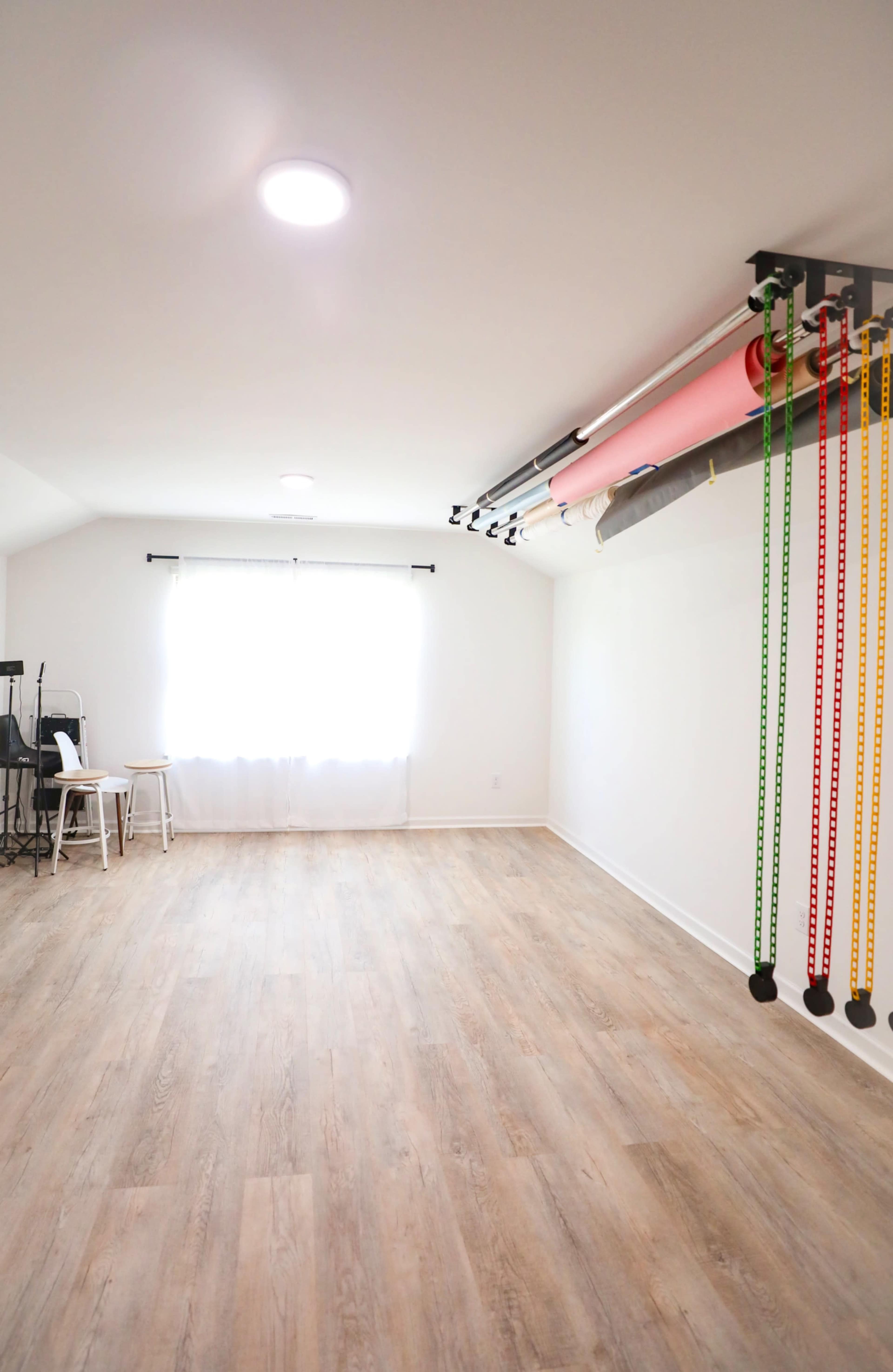 The image shows a spacious, empty room with light wooden flooring, a window with white curtains, and various colorful rolls and resistance bands stored on a mounted rack.