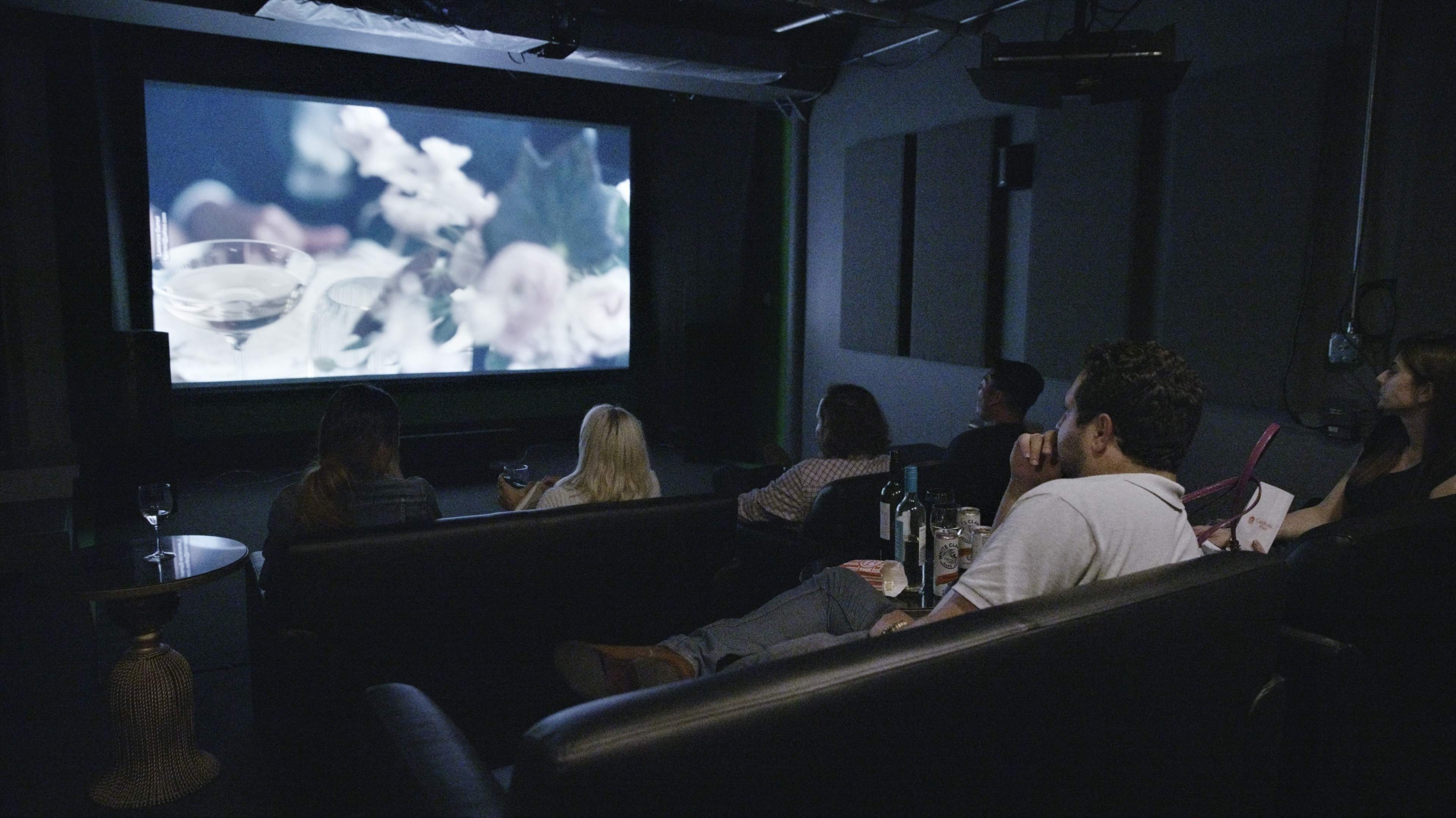 A group of people sits on a couch watching a movie on a large screen in a dimly lit room.