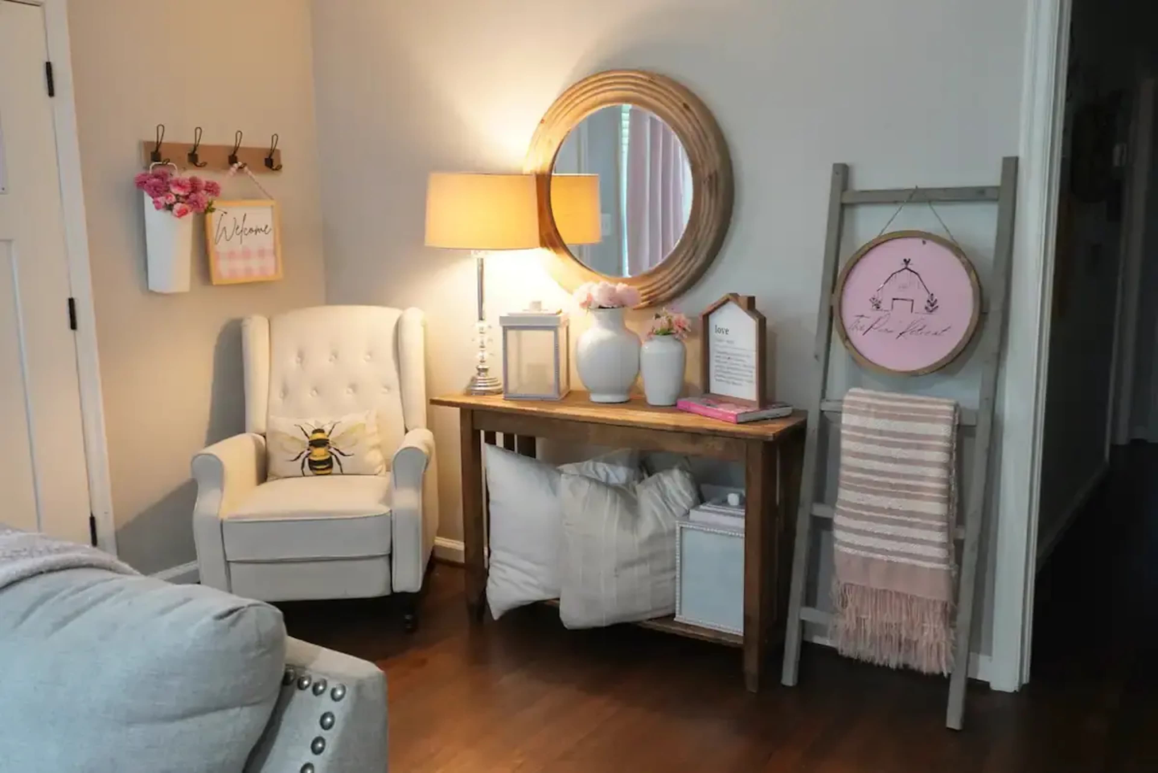 A cozy corner of a room features a white armchair beside a wooden console table, adorned with decorative items and a round mirror, with a ladder displaying a woven blanket.