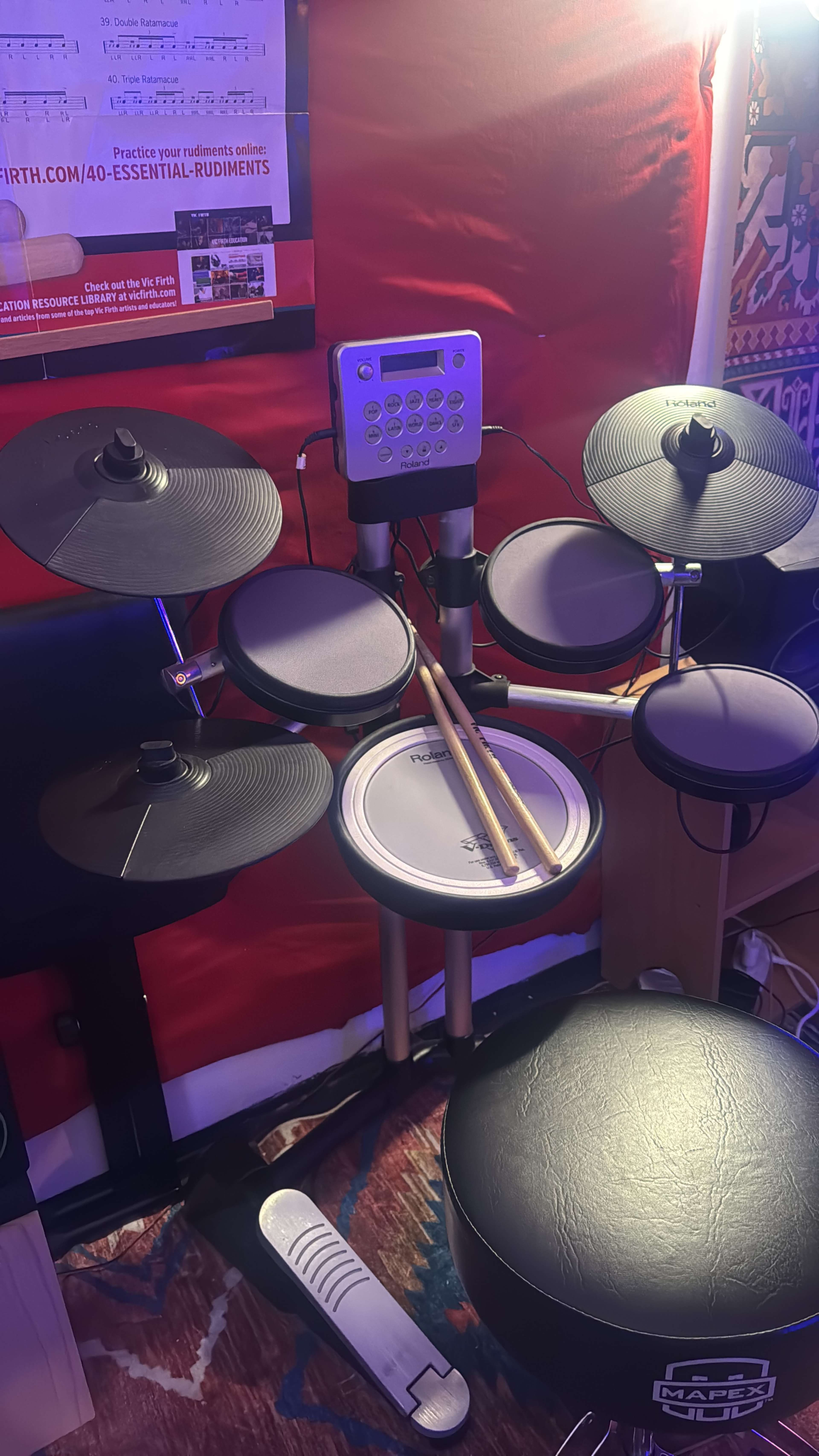 The image shows an electronic drum set with drum pads, a drum throne, and a digital drum machine, situated against a red backdrop.