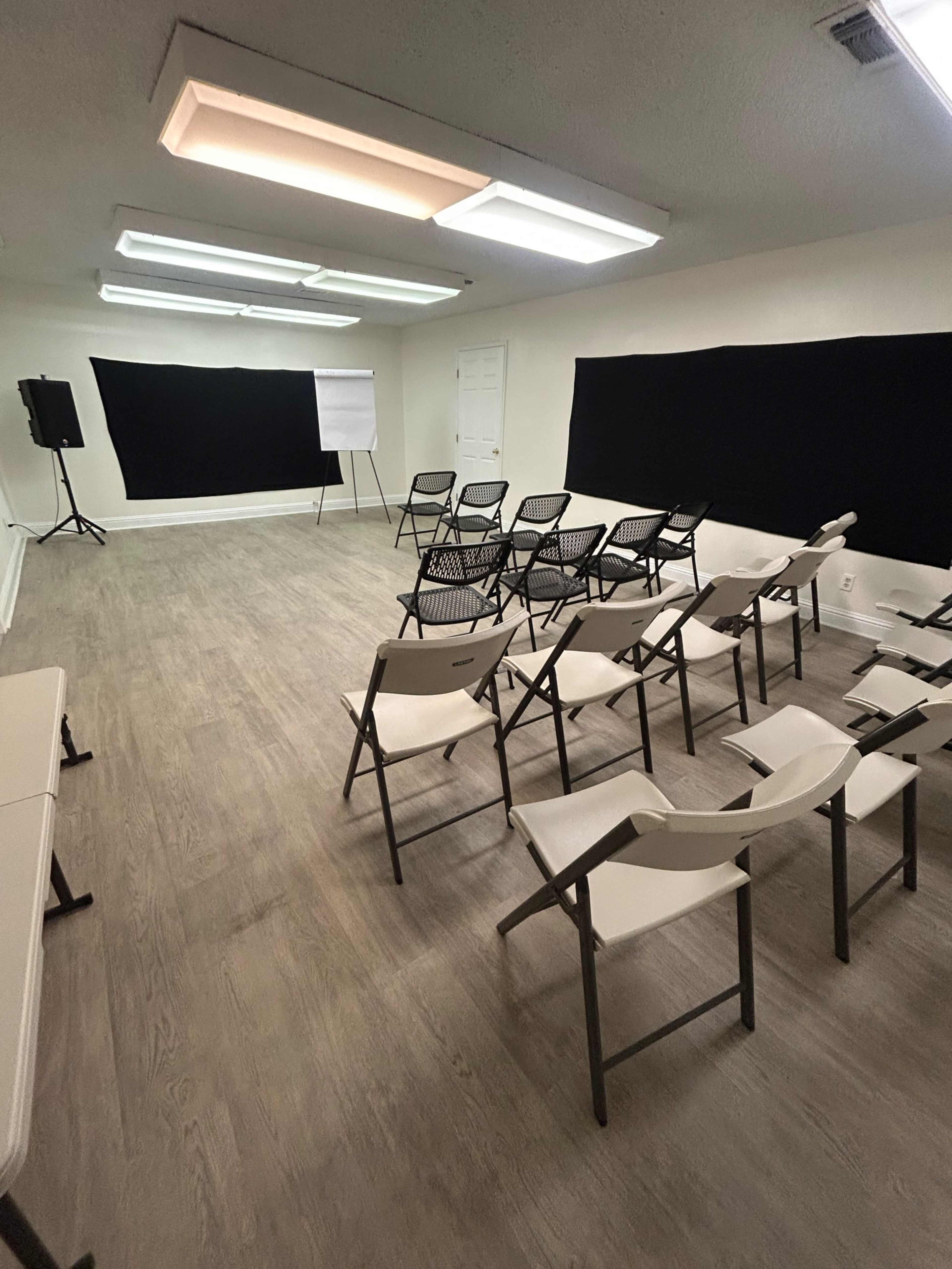 The image shows a sparsely furnished meeting room with rows of folding chairs facing a blank black backdrop, along with a presentation board and some tables along the side.