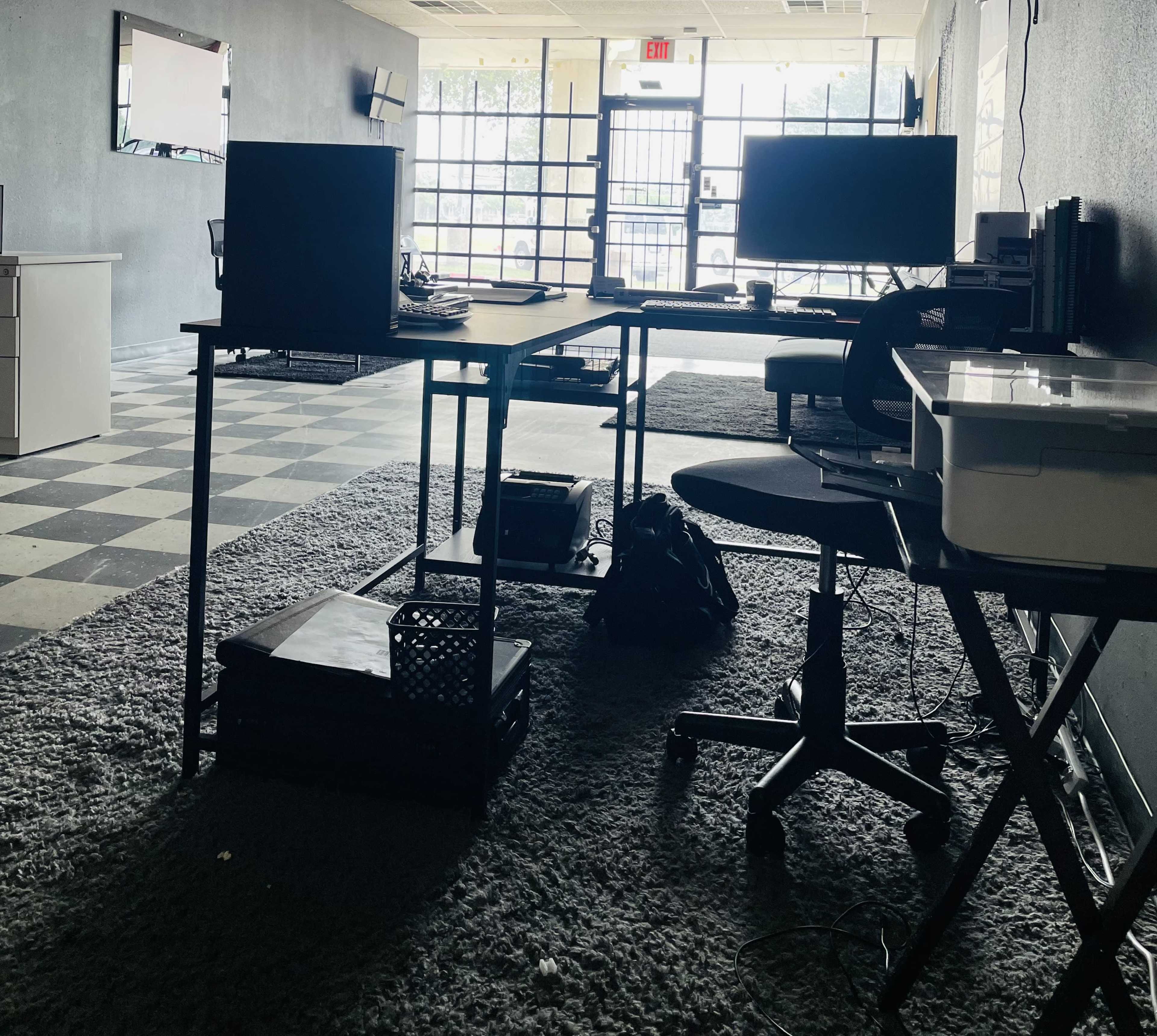 An empty office space with black walls, a checkered floor, and two desks positioned near large windows.