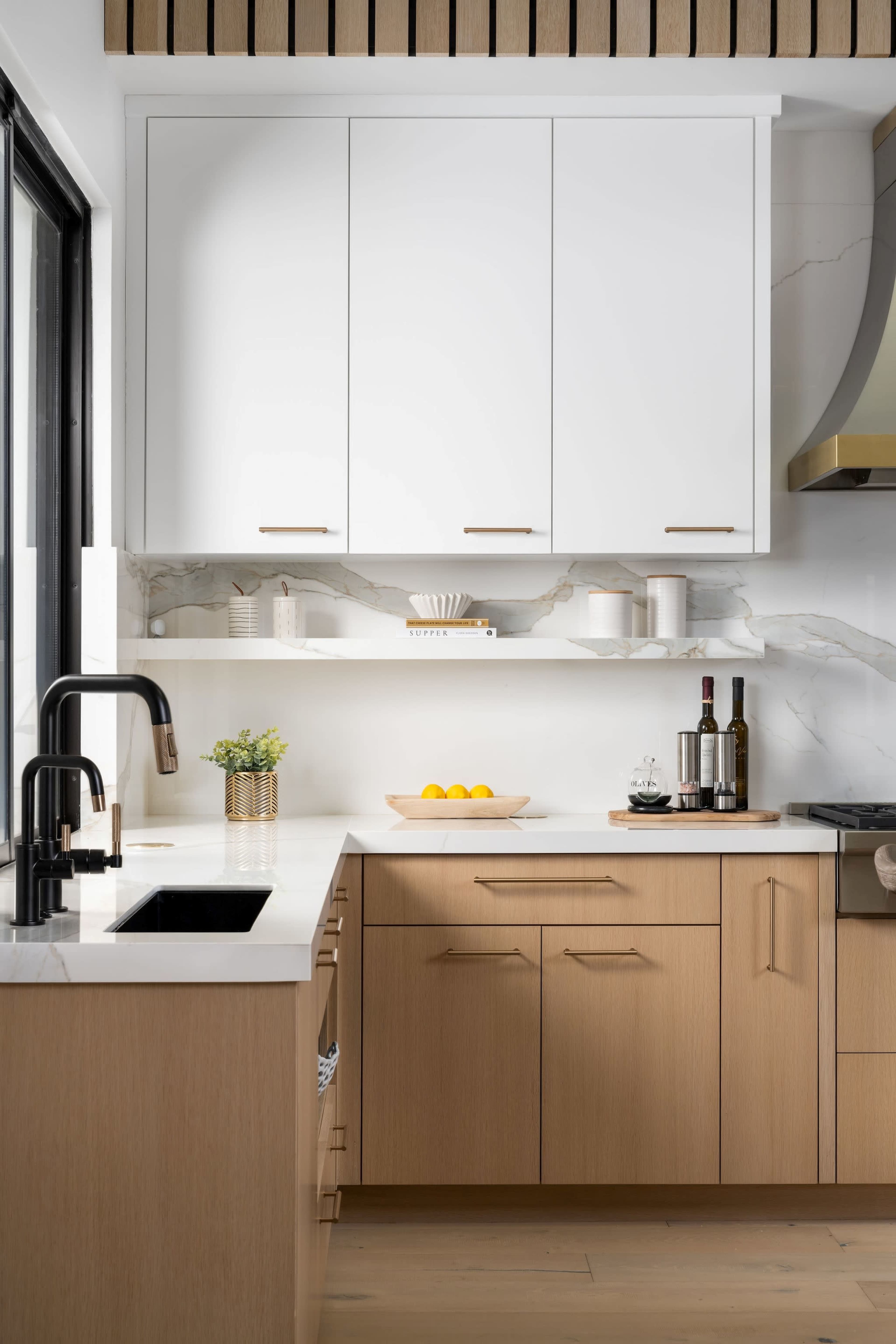 The image shows a modern kitchen with wood and white cabinetry, a black faucet, a marble backsplash, and a countertop displaying fresh fruit and wine.