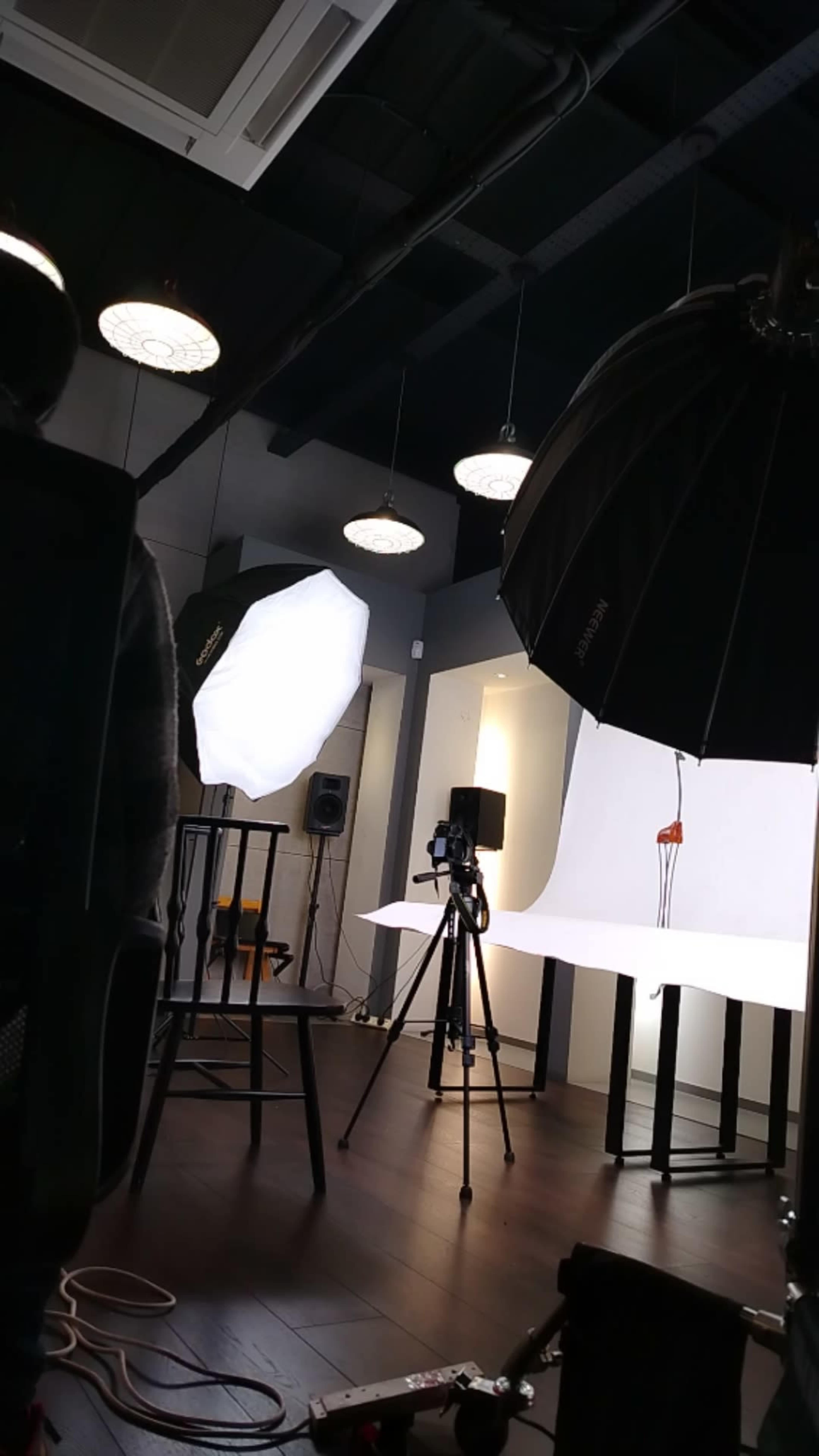 The image depicts a photography studio setup with lighting umbrellas, a camera on a tripod, and a plain backdrop.