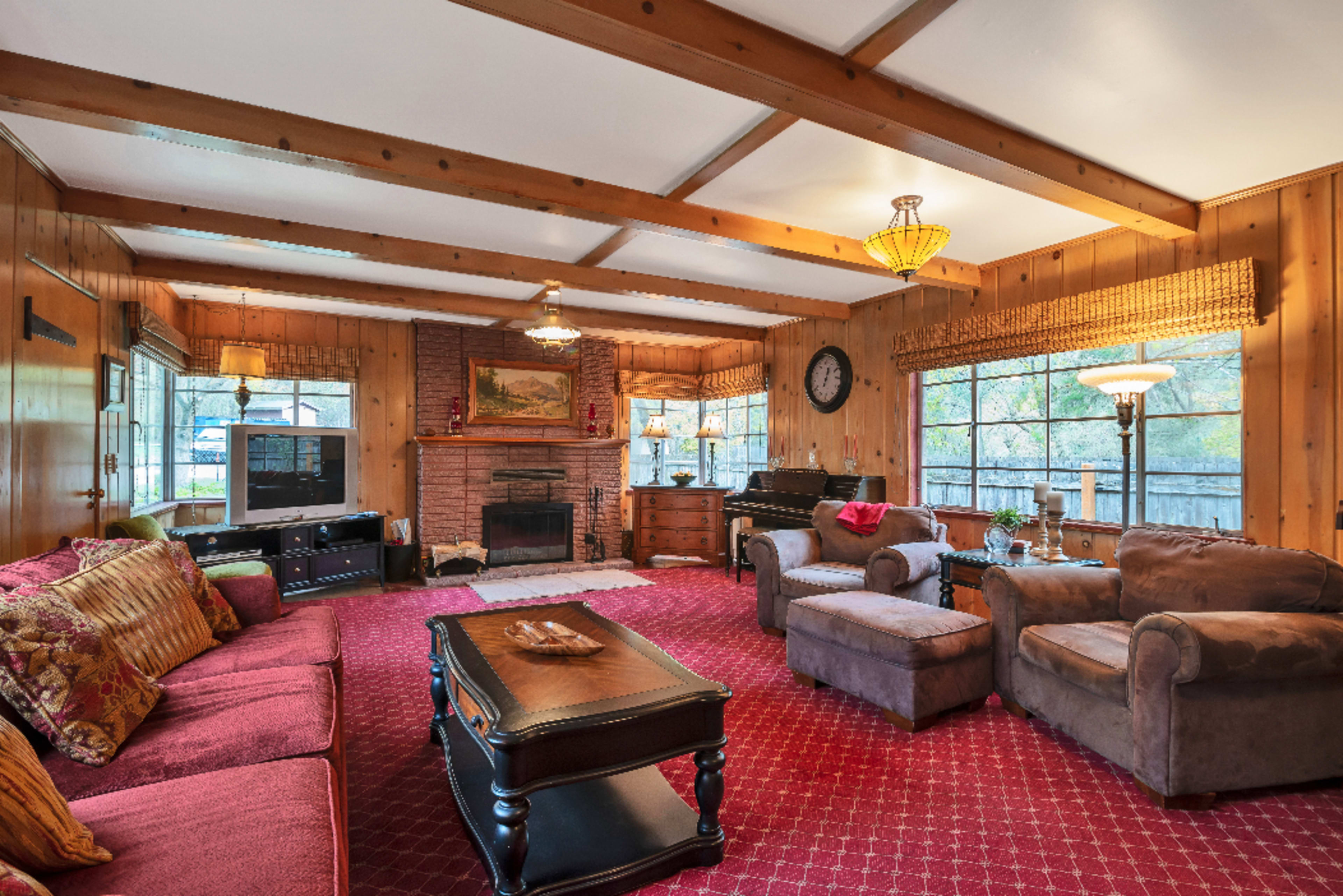 A living room with wooden paneling, a brick fireplace, and multiple seating arrangements, including a couch and armchairs, set on a red carpet.