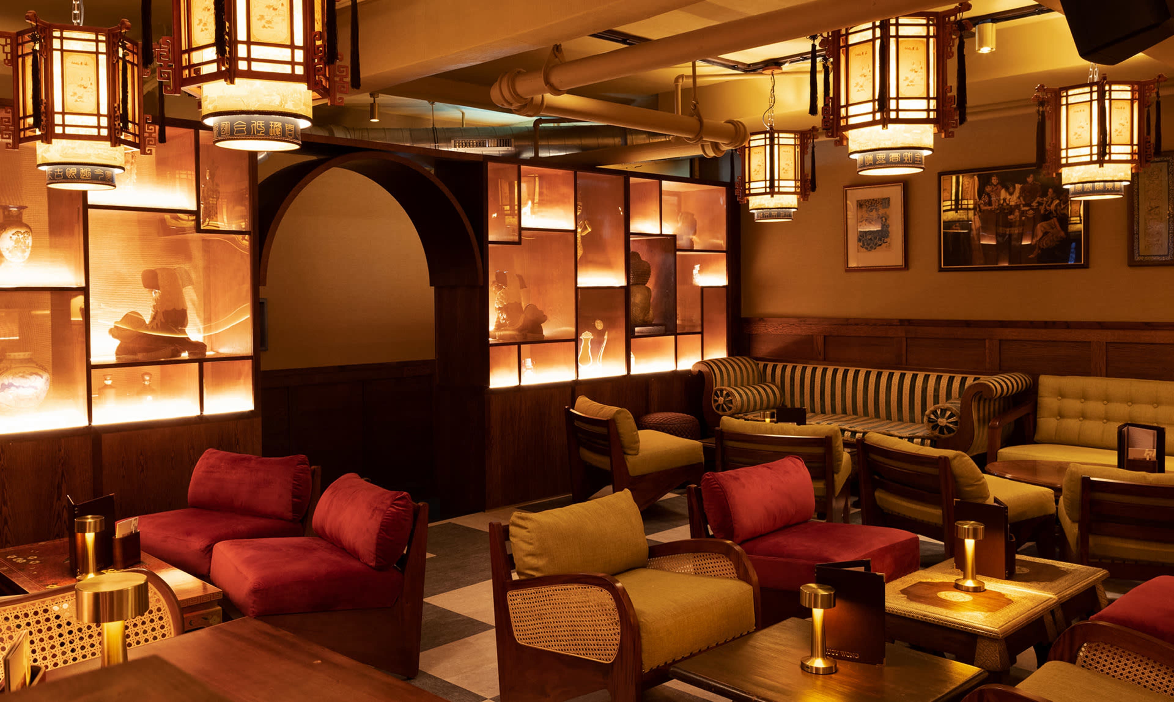 The image shows a warmly lit lounge with plush seating arrangements and decorative lighting fixtures, featuring an archway and illuminated display cases against the walls.