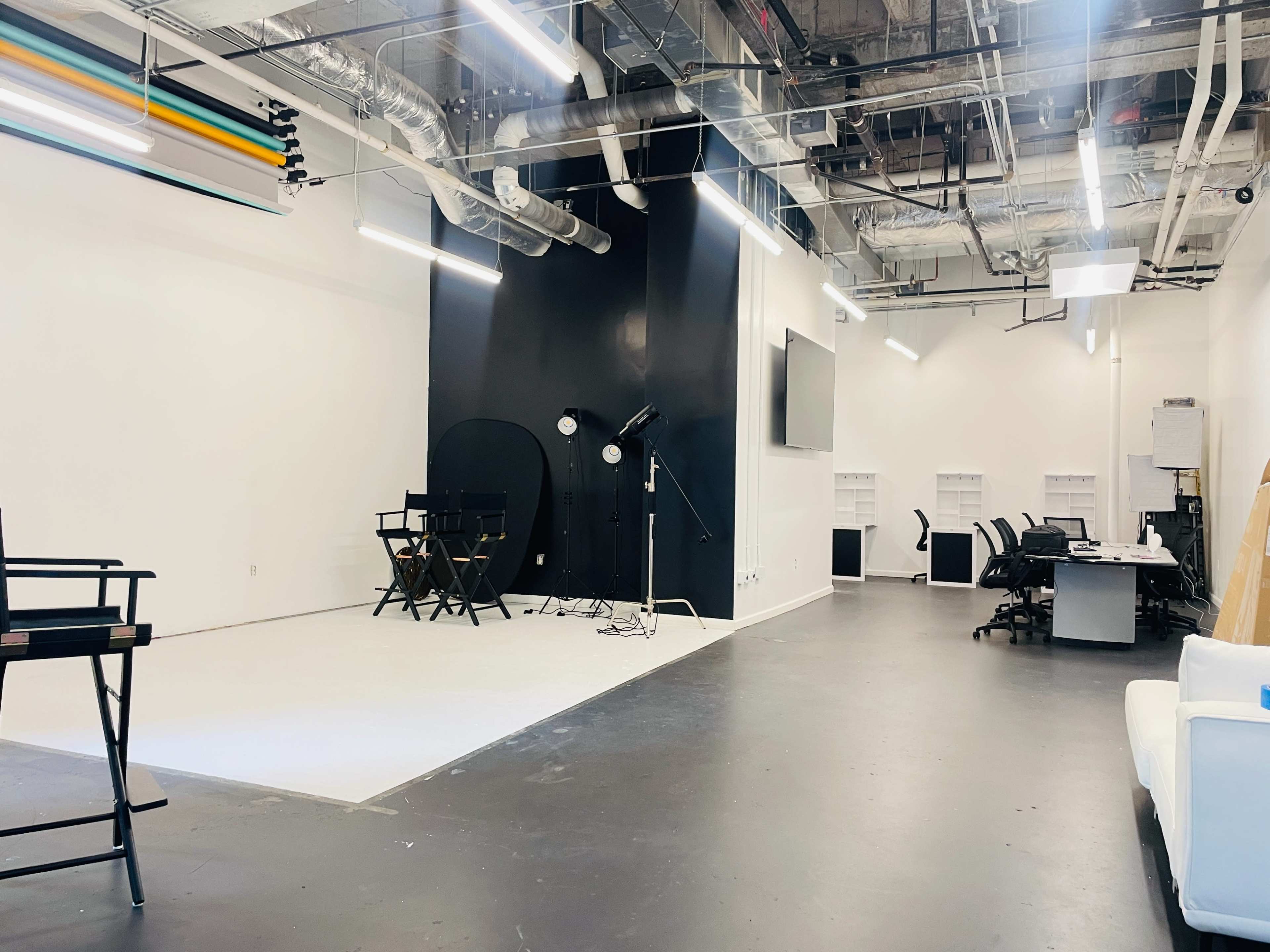 The image shows a spacious, modern studio space with black and white walls, lighting equipment, director's chairs, and a row of desks on one side.
