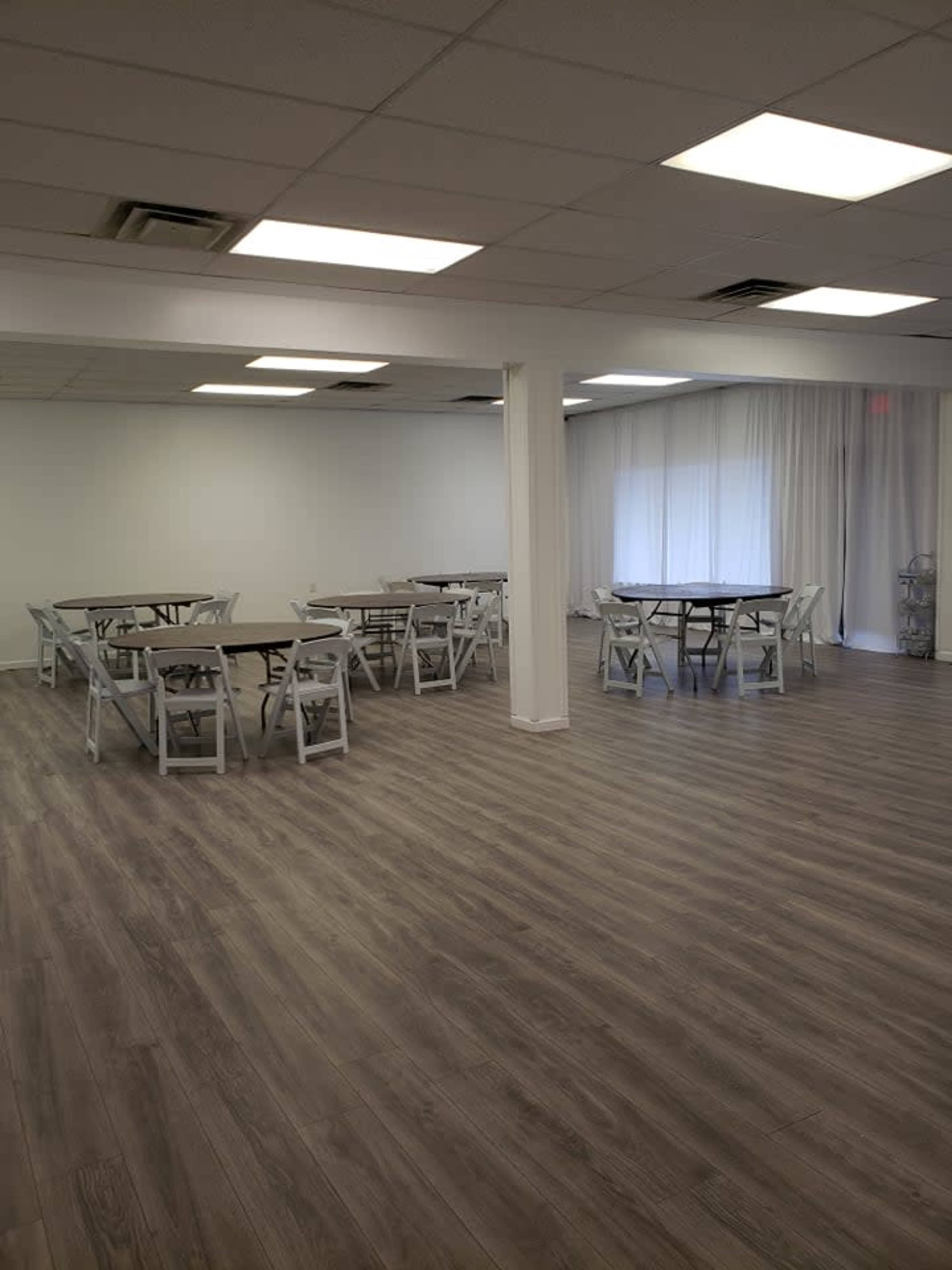 An empty room with gray wooden floors, several round tables surrounded by white folding chairs, and large windows covered by white curtains.