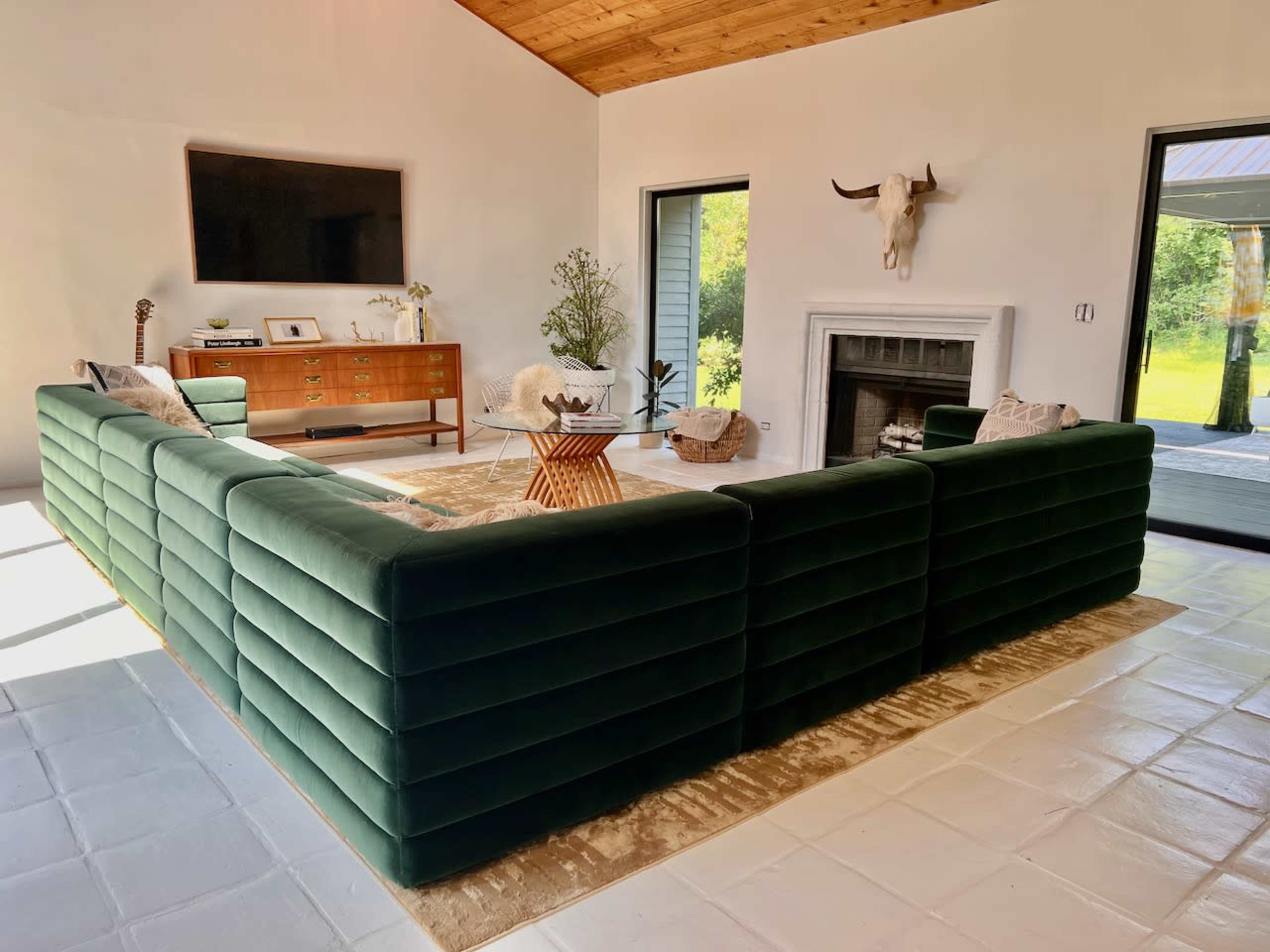 A modern living room features a large green velvet sectional sofa arranged around a central coffee table, with a television mounted on the wall and a fireplace nearby.