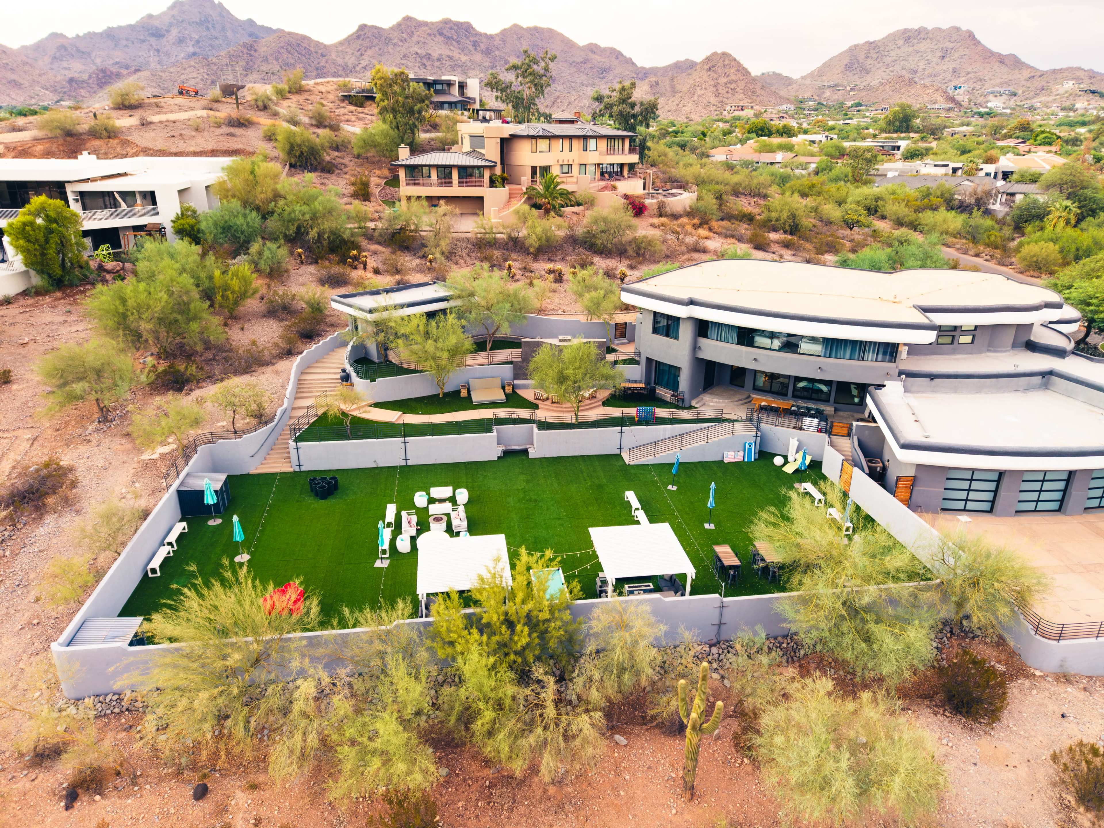 The image shows a modern house situated on a hillside, surrounded by desert vegetation and mountains, with a well-maintained green lawn and outdoor furniture in the yard.