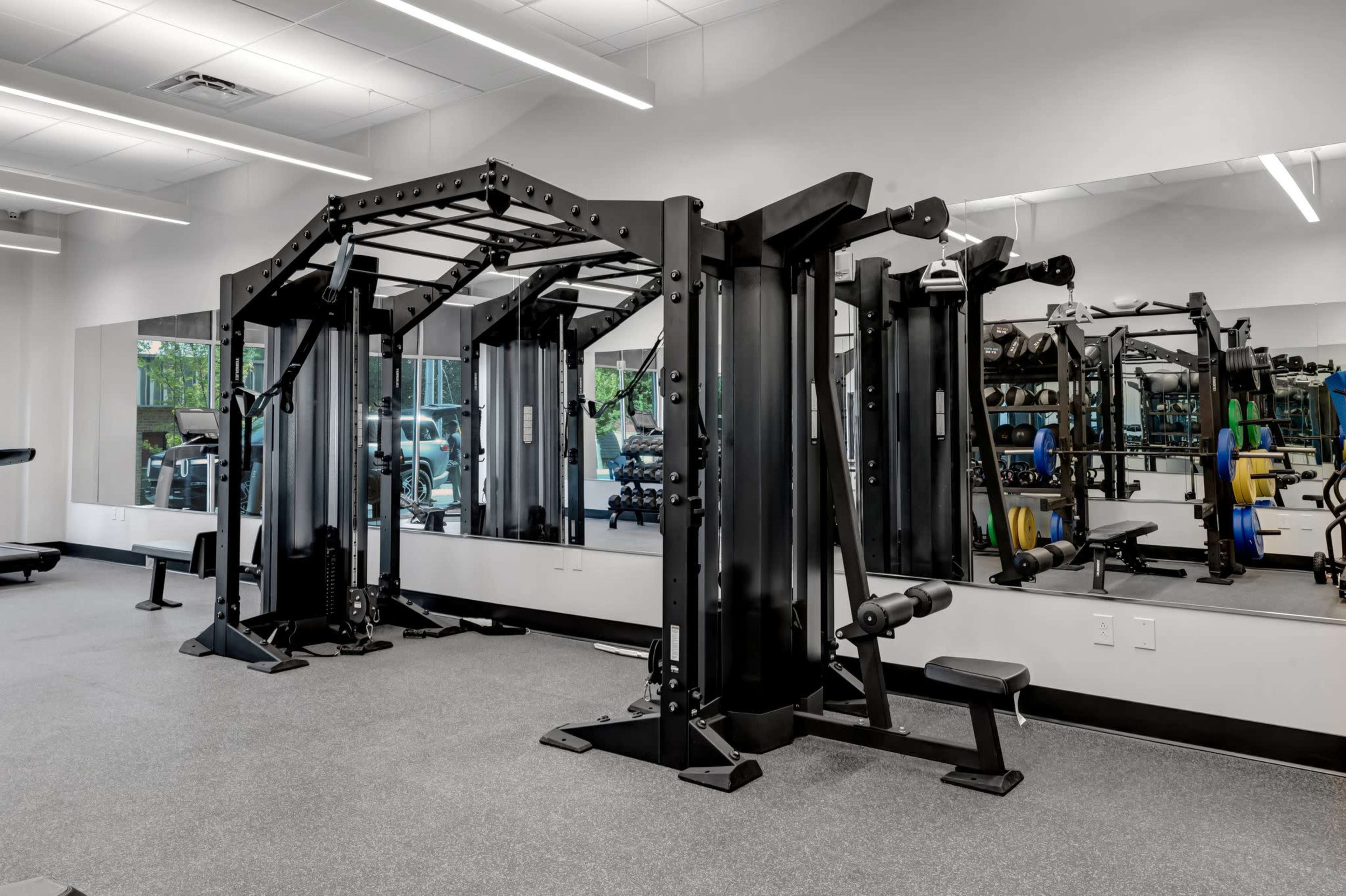 The image shows a well-equipped fitness gym featuring a black multi-station strength training machine and various free weights arranged on multiple racks.