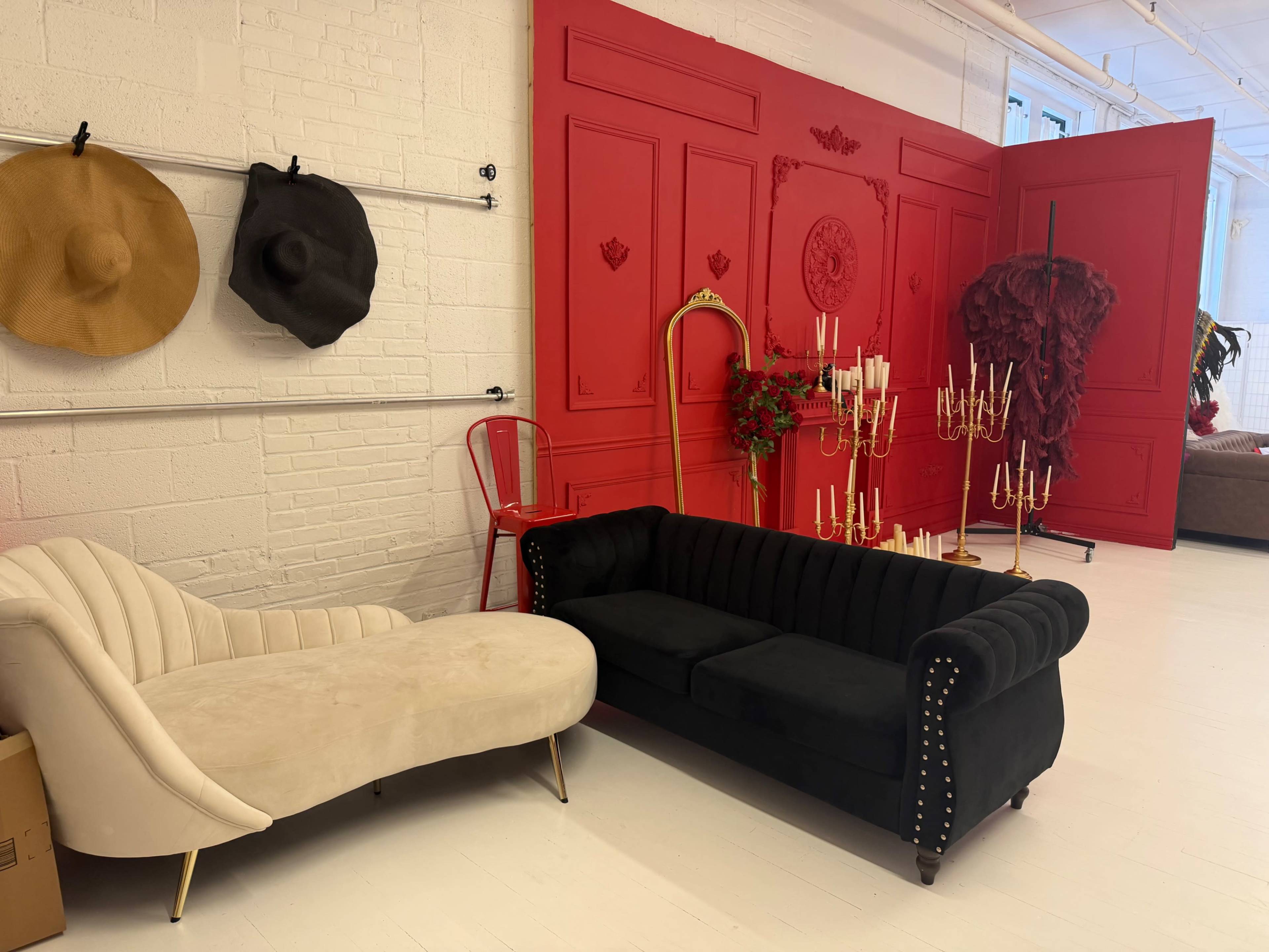 The image shows a stylish interior with a black velvet couch and a beige curved lounge chair against a bright red wall adorned with decorative panels and accessories.