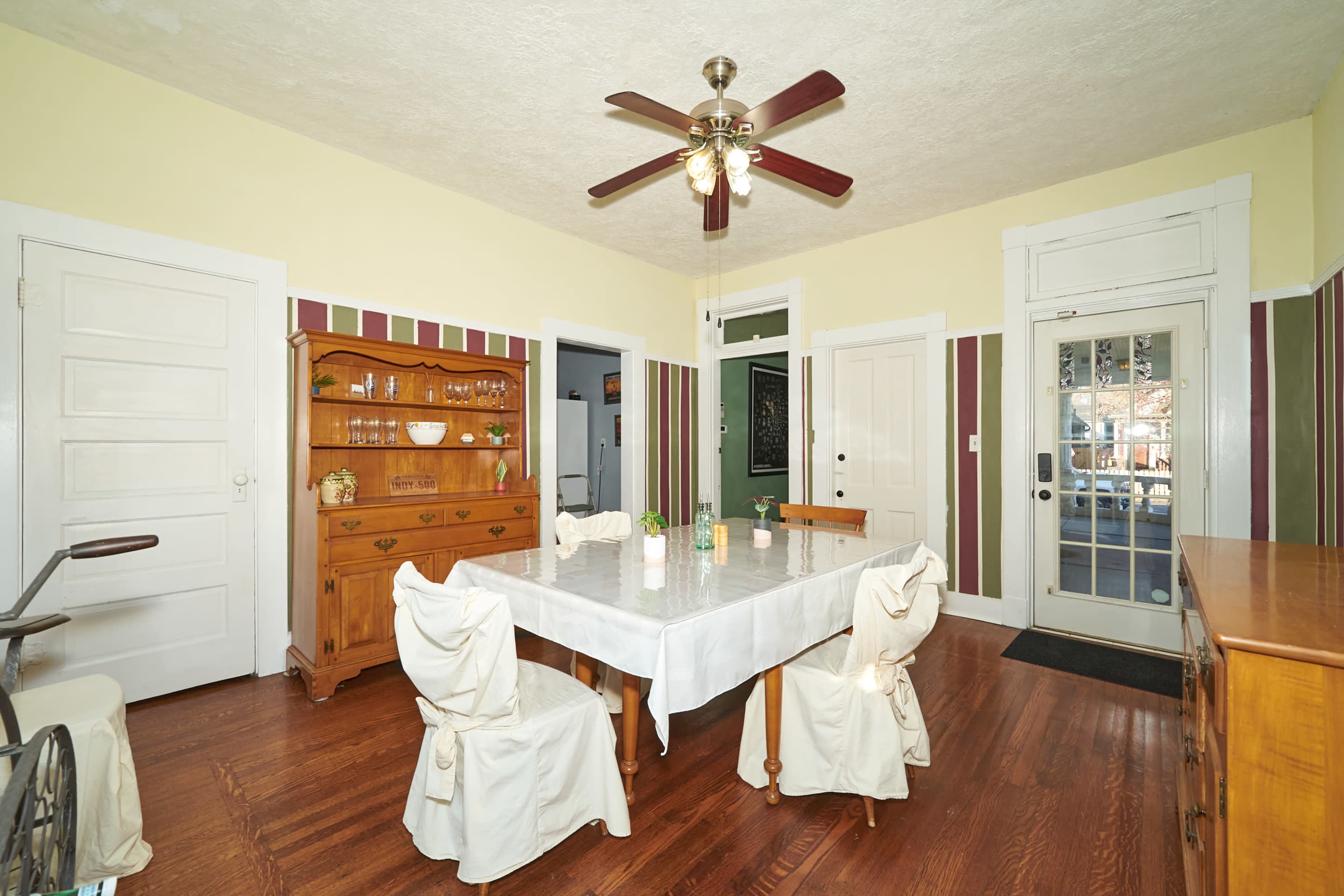 A dining room features a central table surrounded by six chairs, with a wooden sideboard and a ceiling fan above.