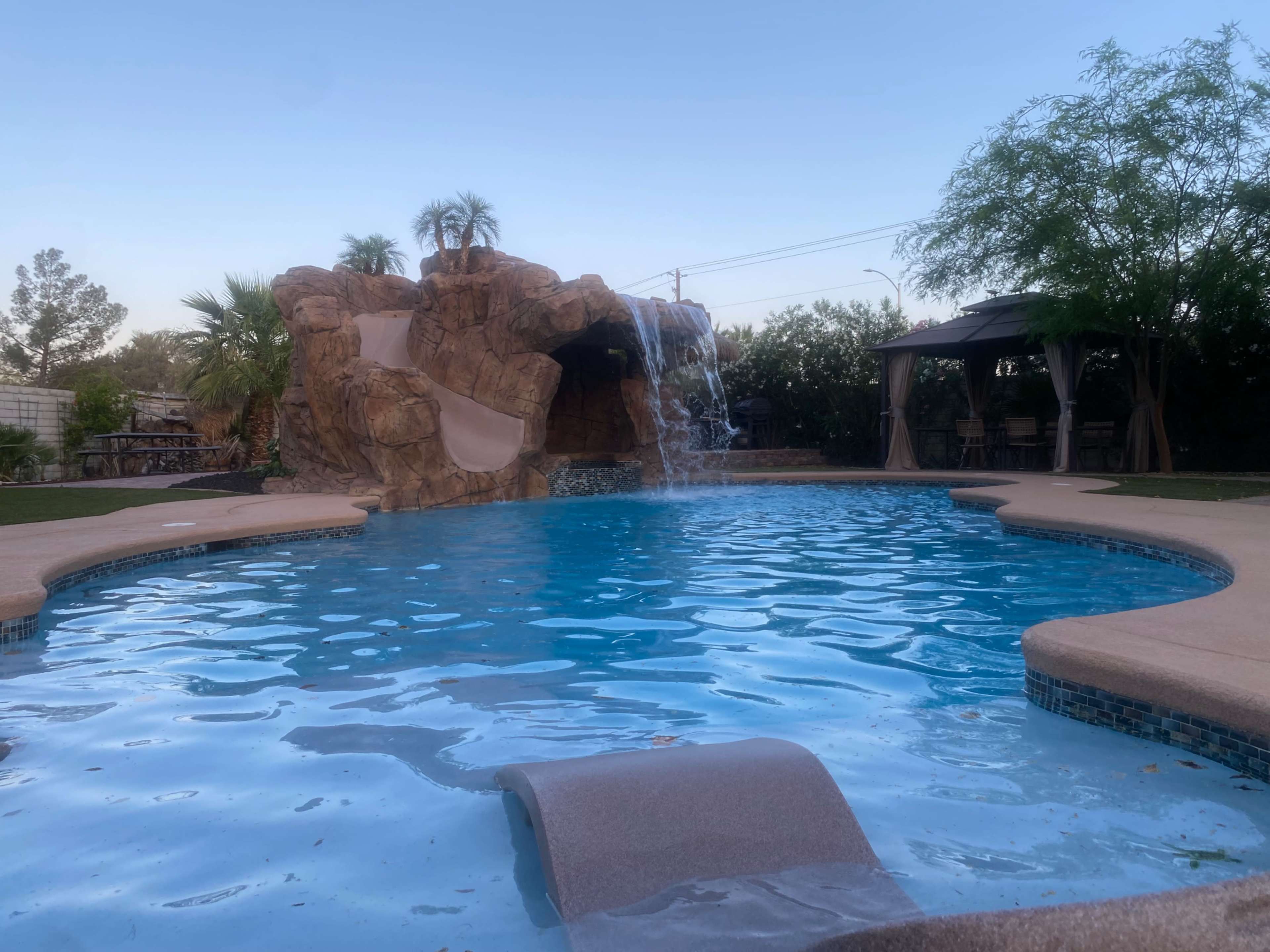 A swimming pool features a rock formation with a waterfall and surrounding palm trees.