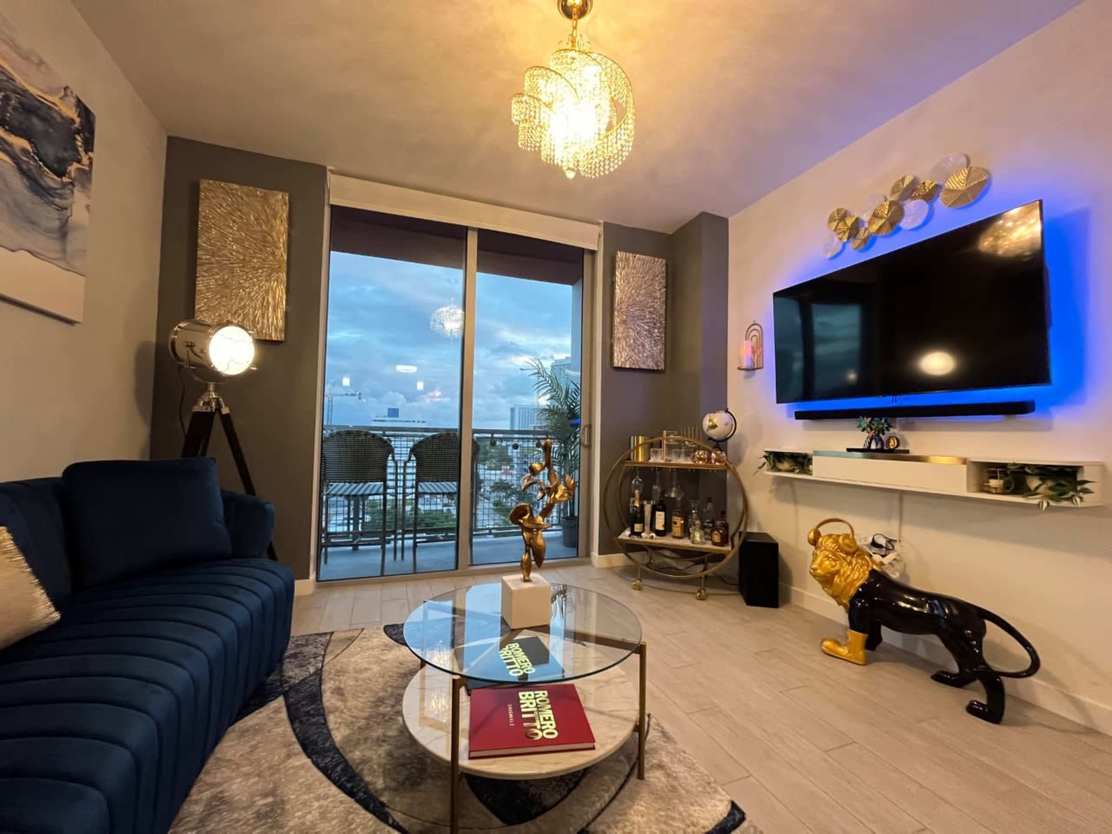 The image shows a modern living room featuring a blue sofa, a glass coffee table with decorative items, a large television mounted on the wall, and a balcony view with outdoor seating.