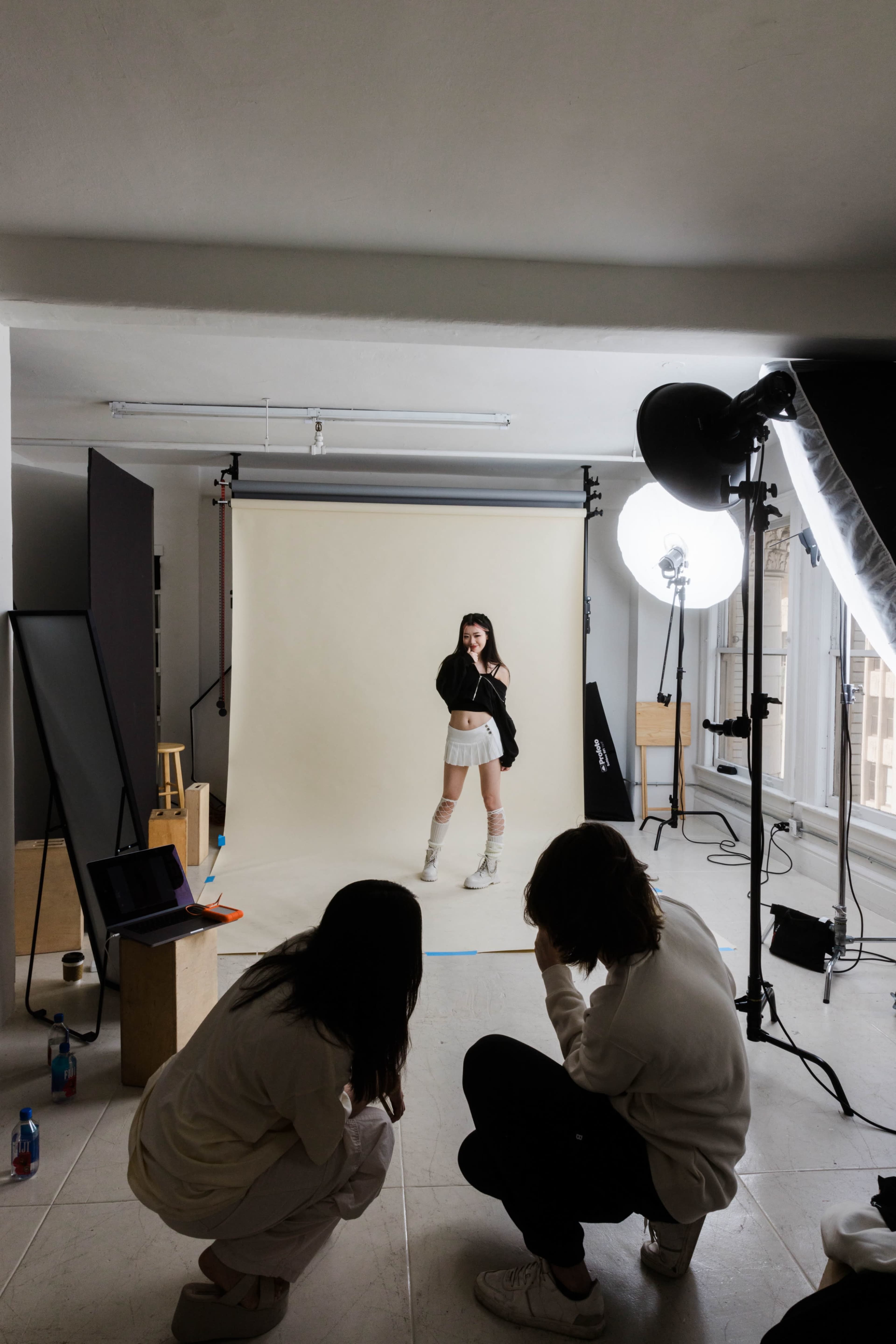 A model poses in front of a neutral backdrop while two photographers observe and adjust their equipment in a studio.
