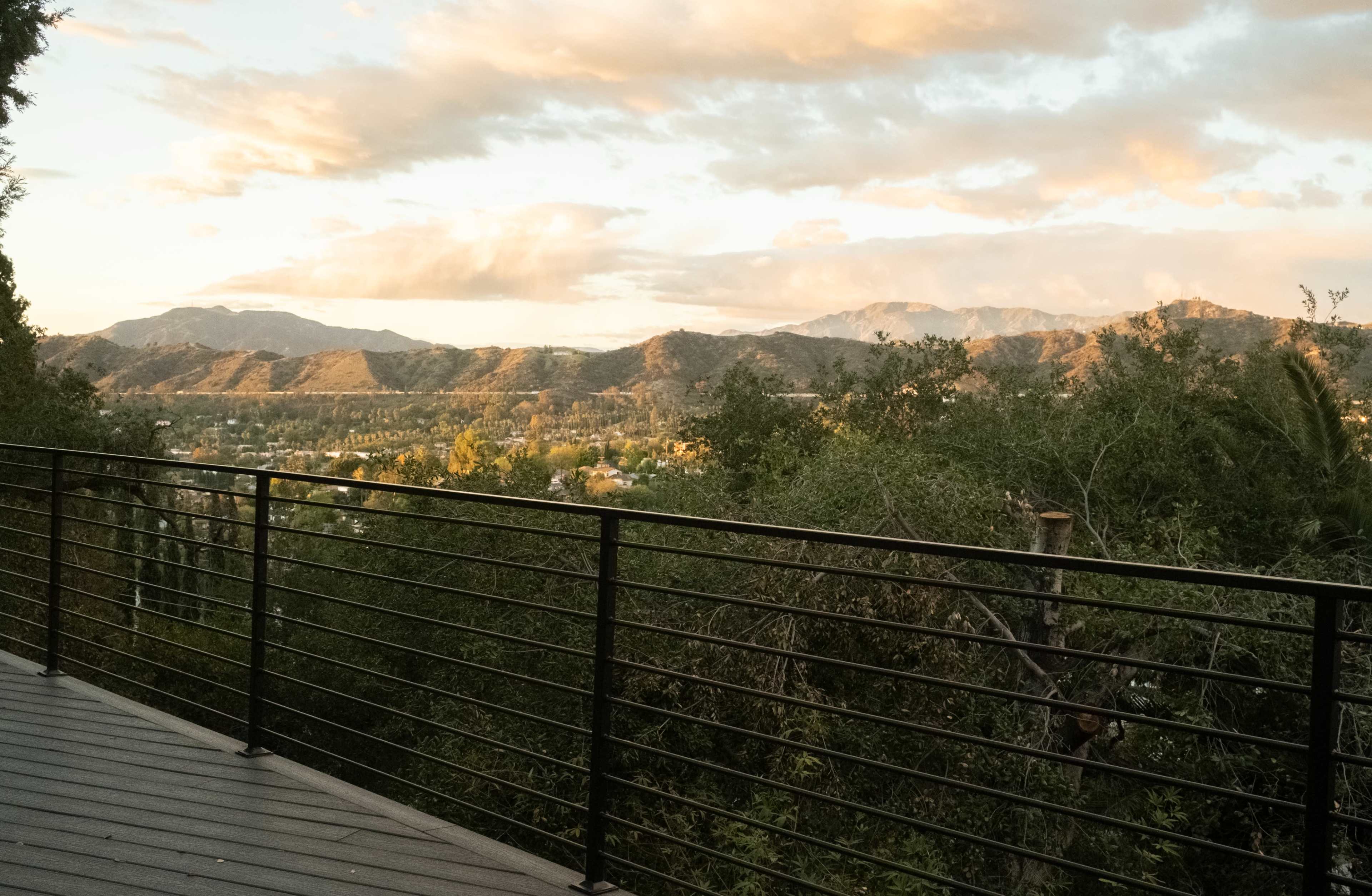 A balcony railing overlooks a valley framed by distant mountains under a partly cloudy sky at sunset.