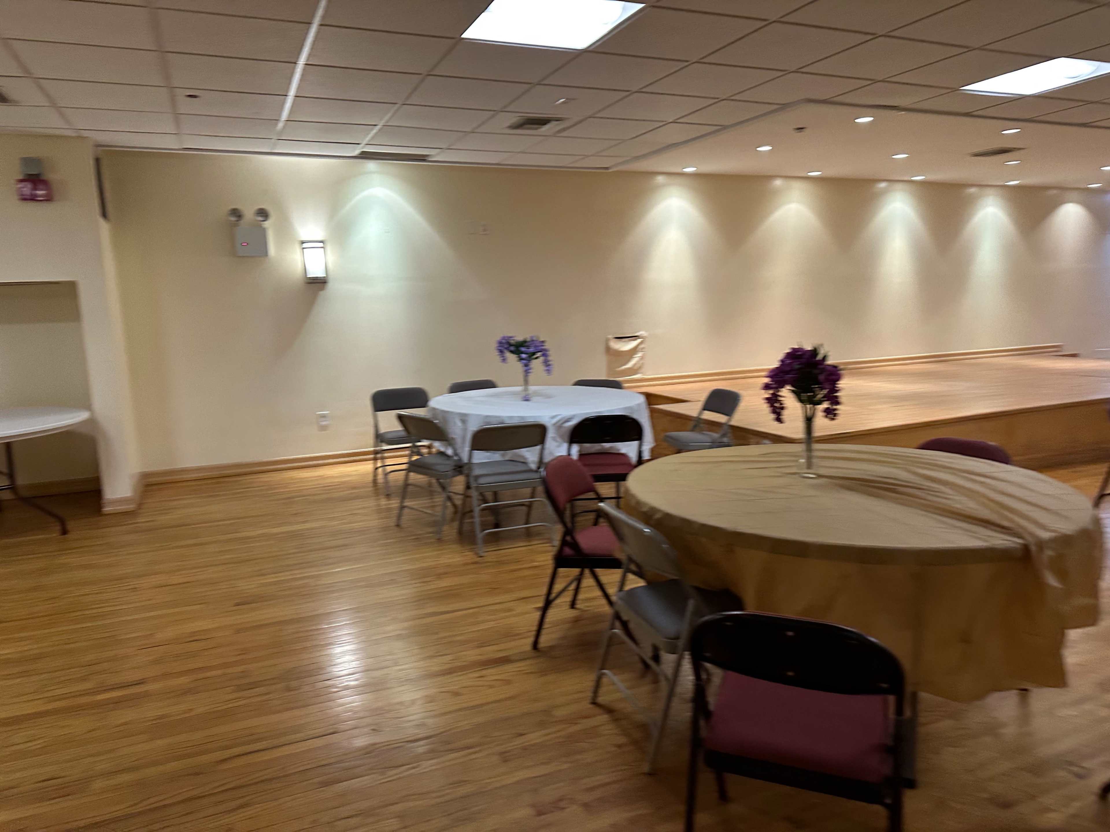 A spacious room with wooden flooring, featuring several tables set with tablecloths and floral centerpieces, and a small stage at one end.
